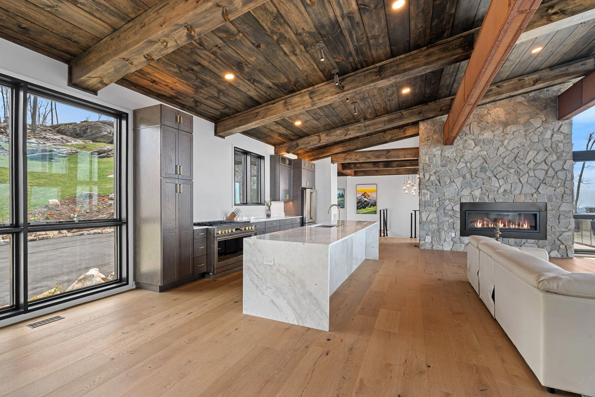 View of a modern kitchen with wooden ceiling beams, stone fireplace, large windows showing outdoor scenery, and an island with a white marble countertop.