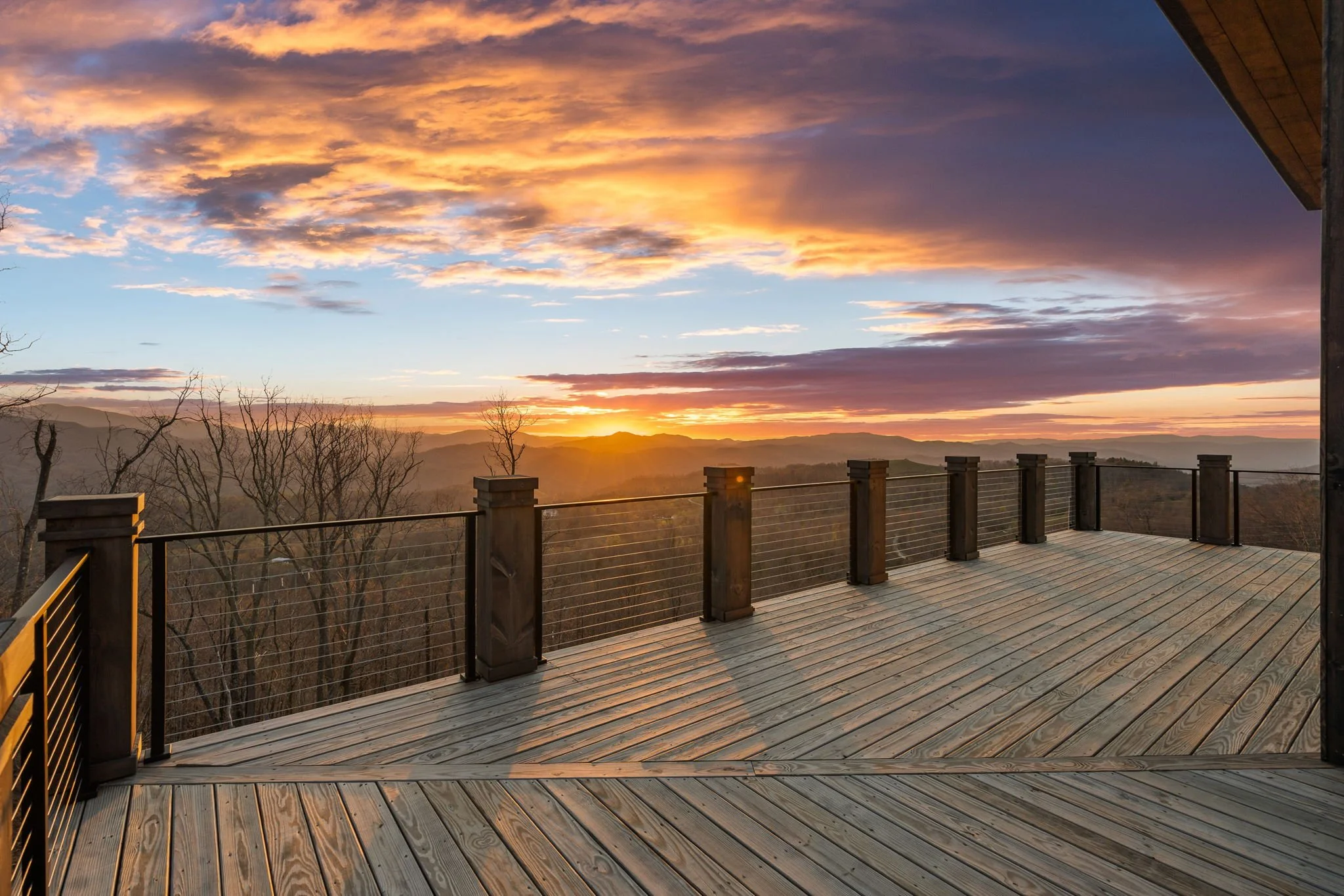 A large outdoor wooden deck with railing overlooking a scenic mountain view during sunset, with clouds in the sky and some bare trees visible in the distance.