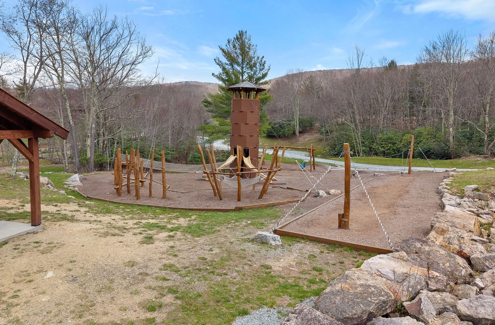 Playground with a wooden climbing structure and slides, surrounded by trees and a mountain in the background.