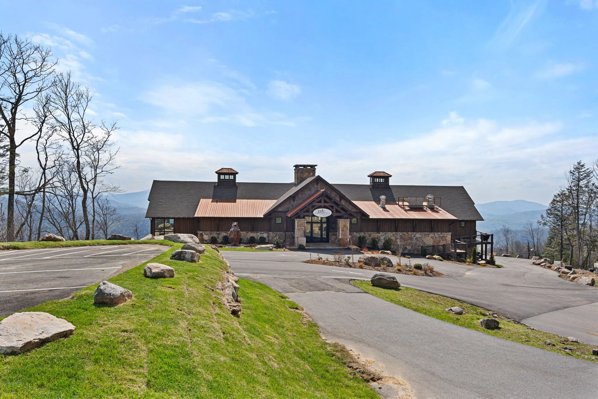 A mountain lodge with a stone and wood exterior, stone chimney, and a sign that reads 'Epic Climbing'. It is surrounded by a parking lot with rocks, patches of grass, and leafless trees, with blue mountains and sky in the background.