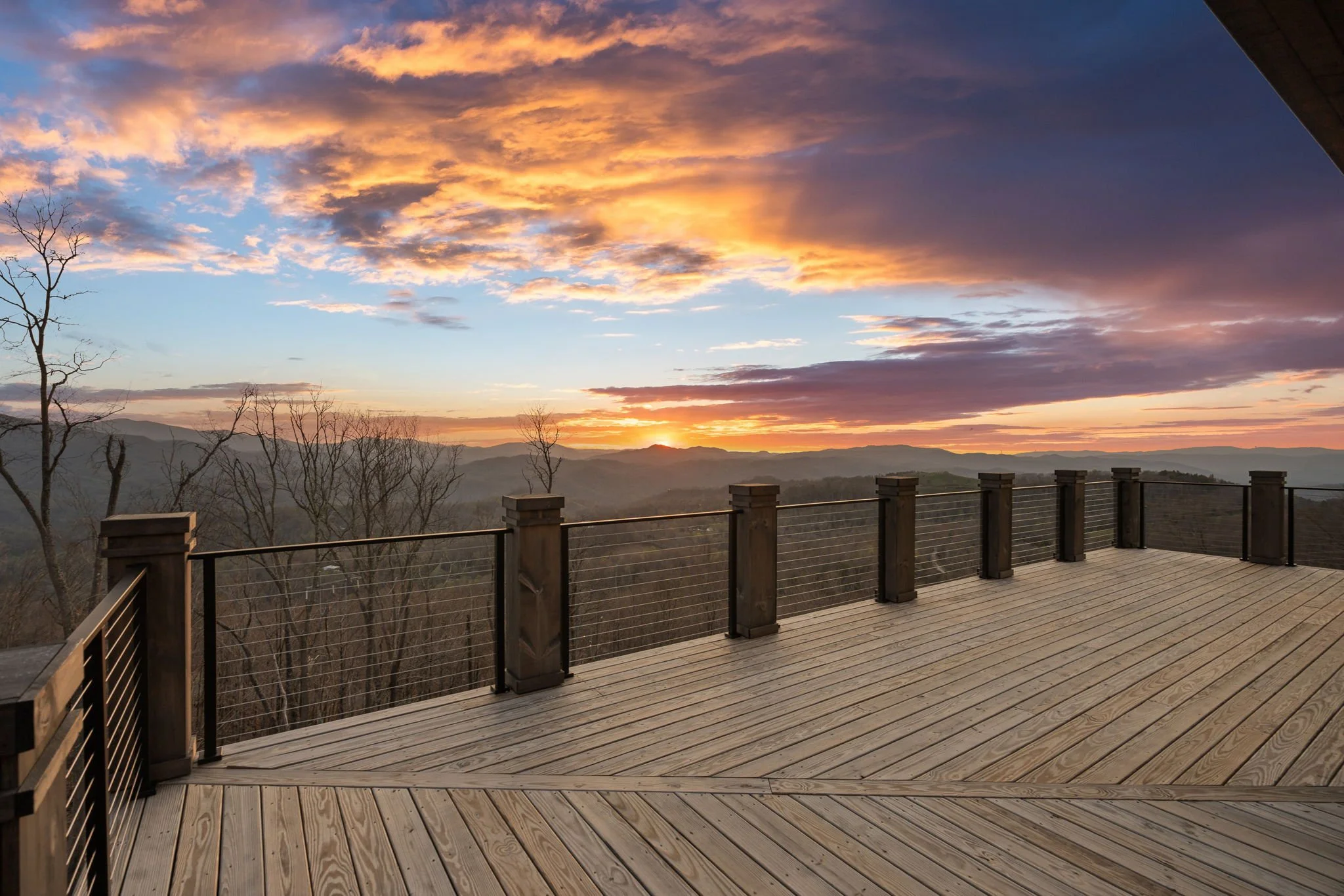 A spacious outdoor wooden deck with a view of hills and mountains at sunset, featuring a railing and leafless trees.