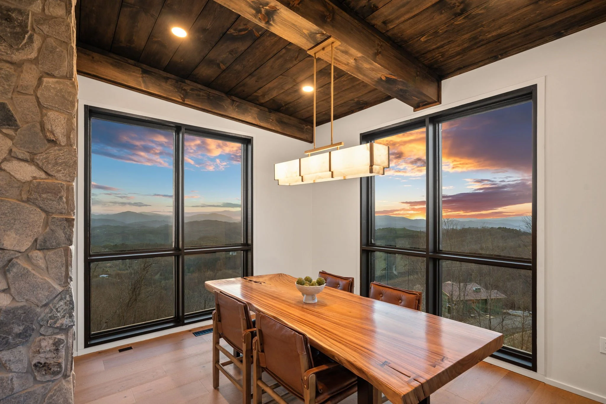 Room with large windows showing a mountain landscape during sunset, wooden ceiling, stone wall, modern light fixture above the dining table, wooden dining table with chairs and a bowl of decorative objects.