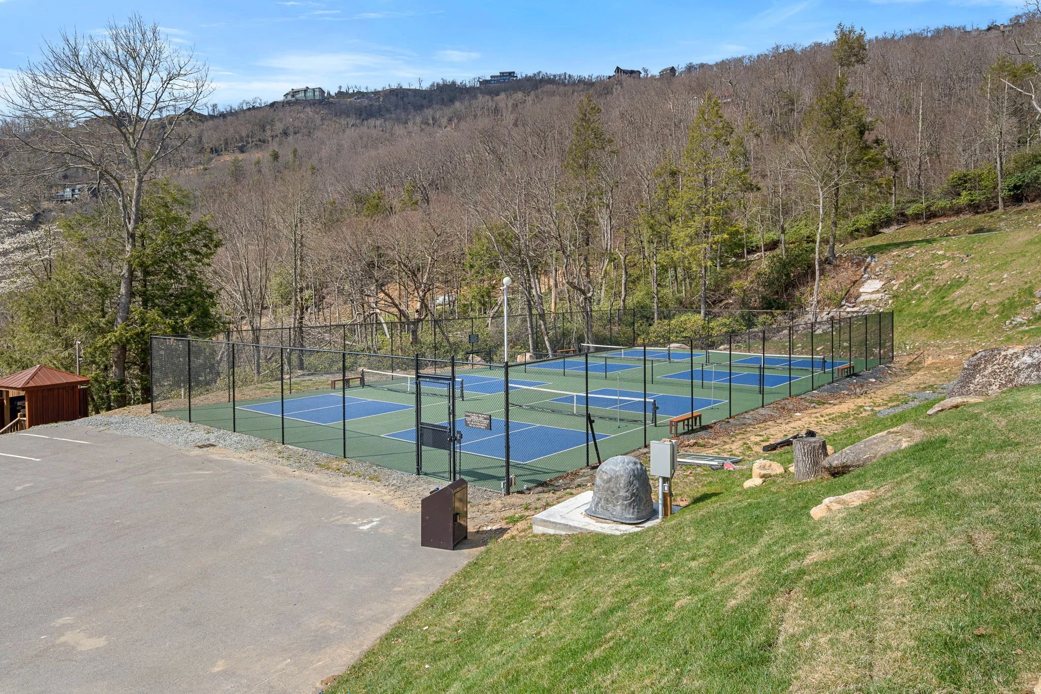 A backyard with a tennis court enclosed by a black fence, surrounded by trees and rocky terrain, with hills and houses in the background under a blue sky.