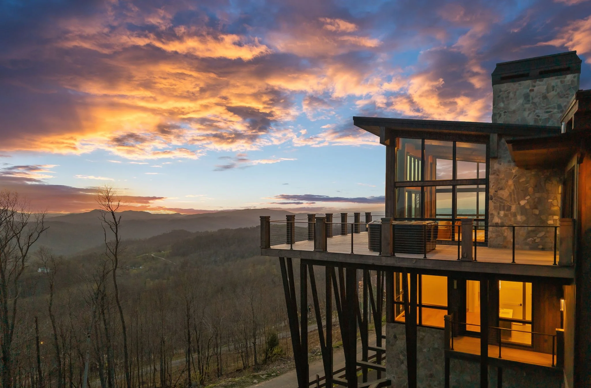 A modern house with large windows and a stone and wood exterior, built on stilts, overlooking a mountainous landscape during sunset with orange and pink clouds in the sky.
