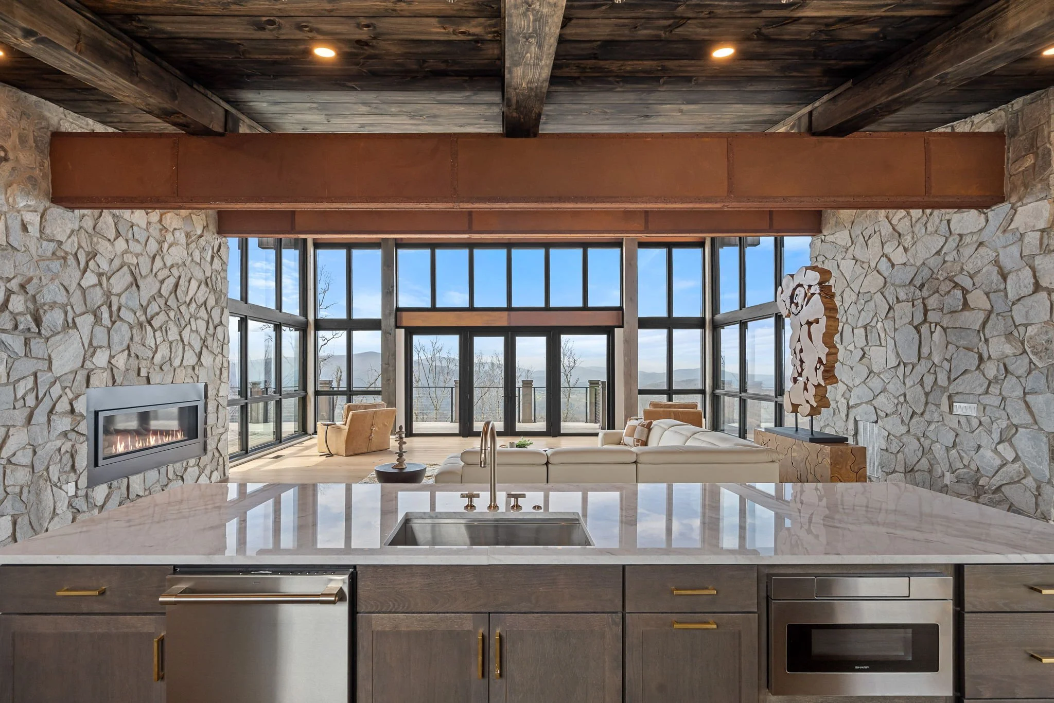 Modern kitchen with a view living room with large windows, stone walls, and a stone fireplace, visible from the kitchen island.
