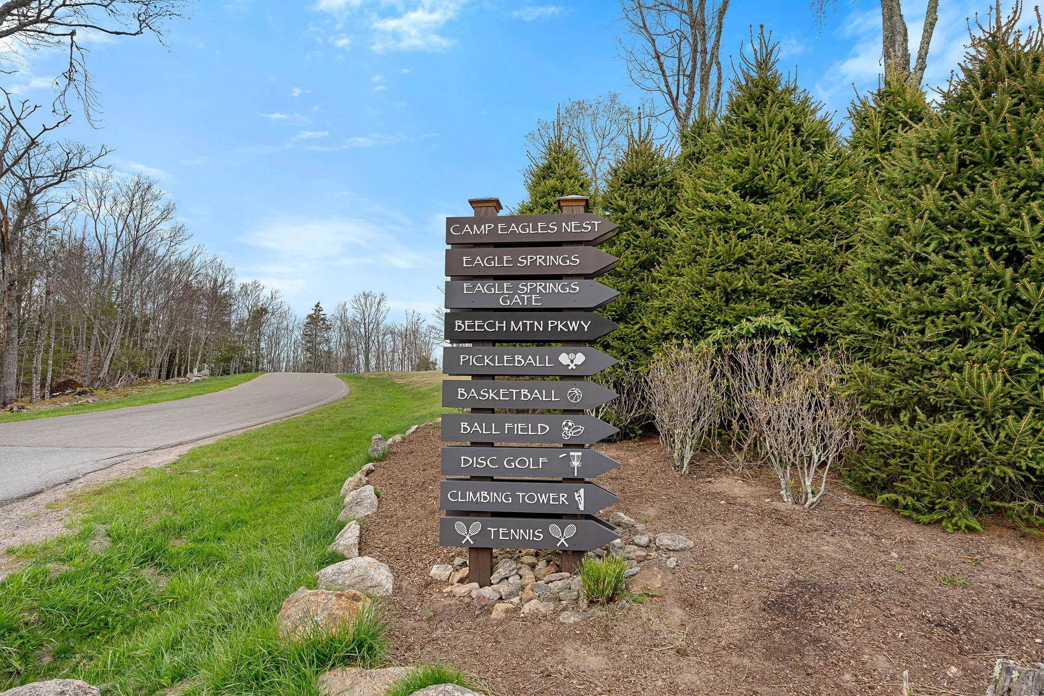 A wooden signpost with black directional signs pointing to various outdoor activity locations, including Camp Eagles Nest, Eagle Springs, Eagle Springs Gate, Beech Mountain Parkway, Pickleball, Basketball, Ball Field, Disc Golf, Climbing Tower, and T