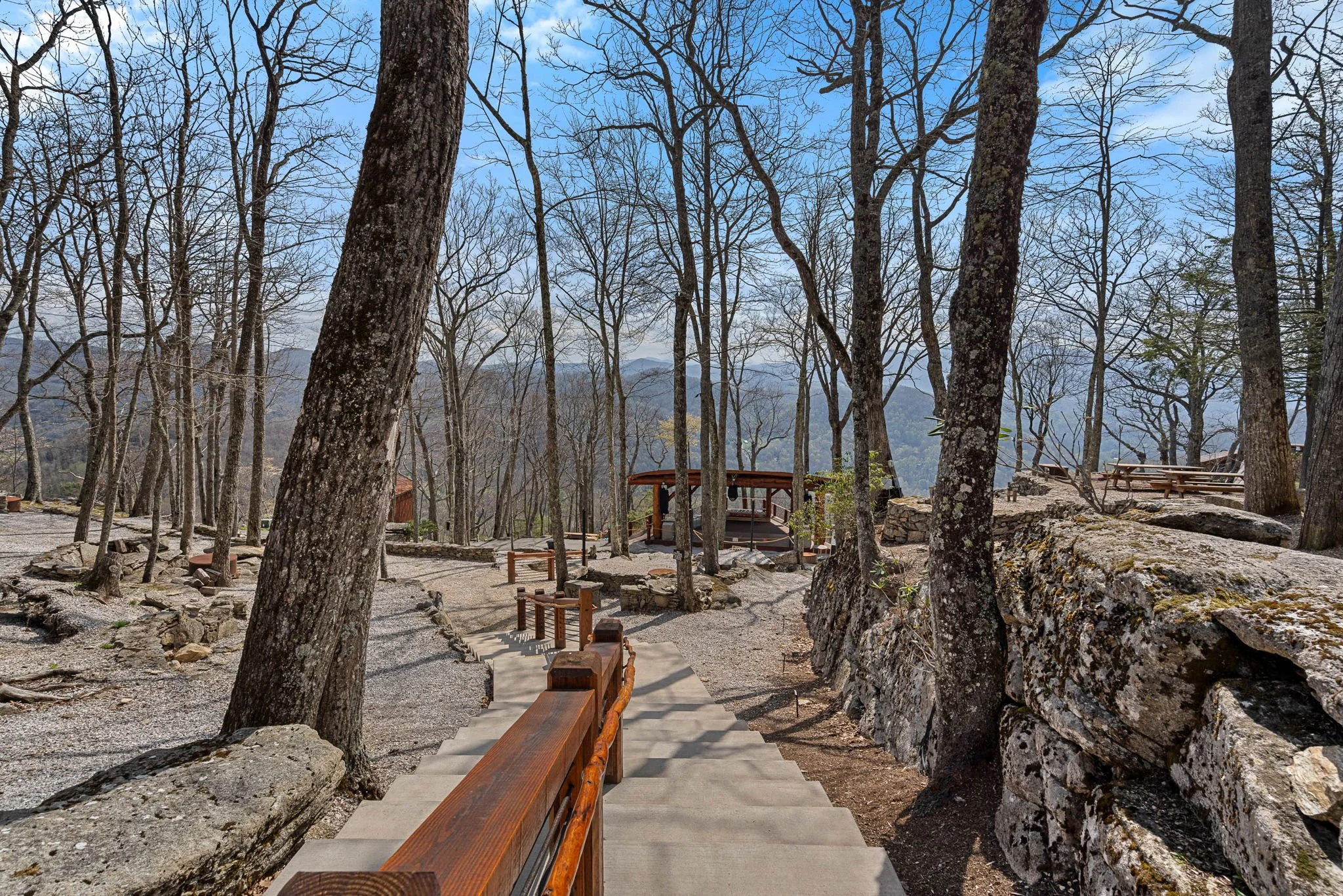 Wooden stairs leading down through a wooded area with leafless trees and rocks, with distant mountains visible in the background under a partly cloudy sky.