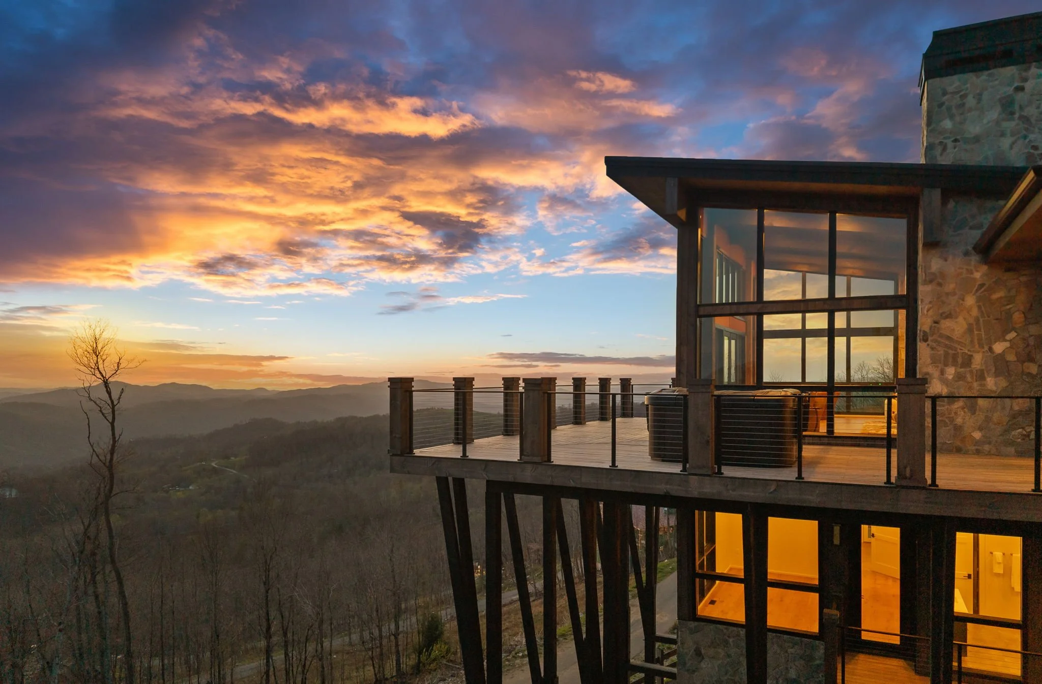A modern house with large glass windows and a stone exterior, perched on a hillside with scenic mountain views at sunset, showing a colorful sky with orange, pink, and blue clouds.