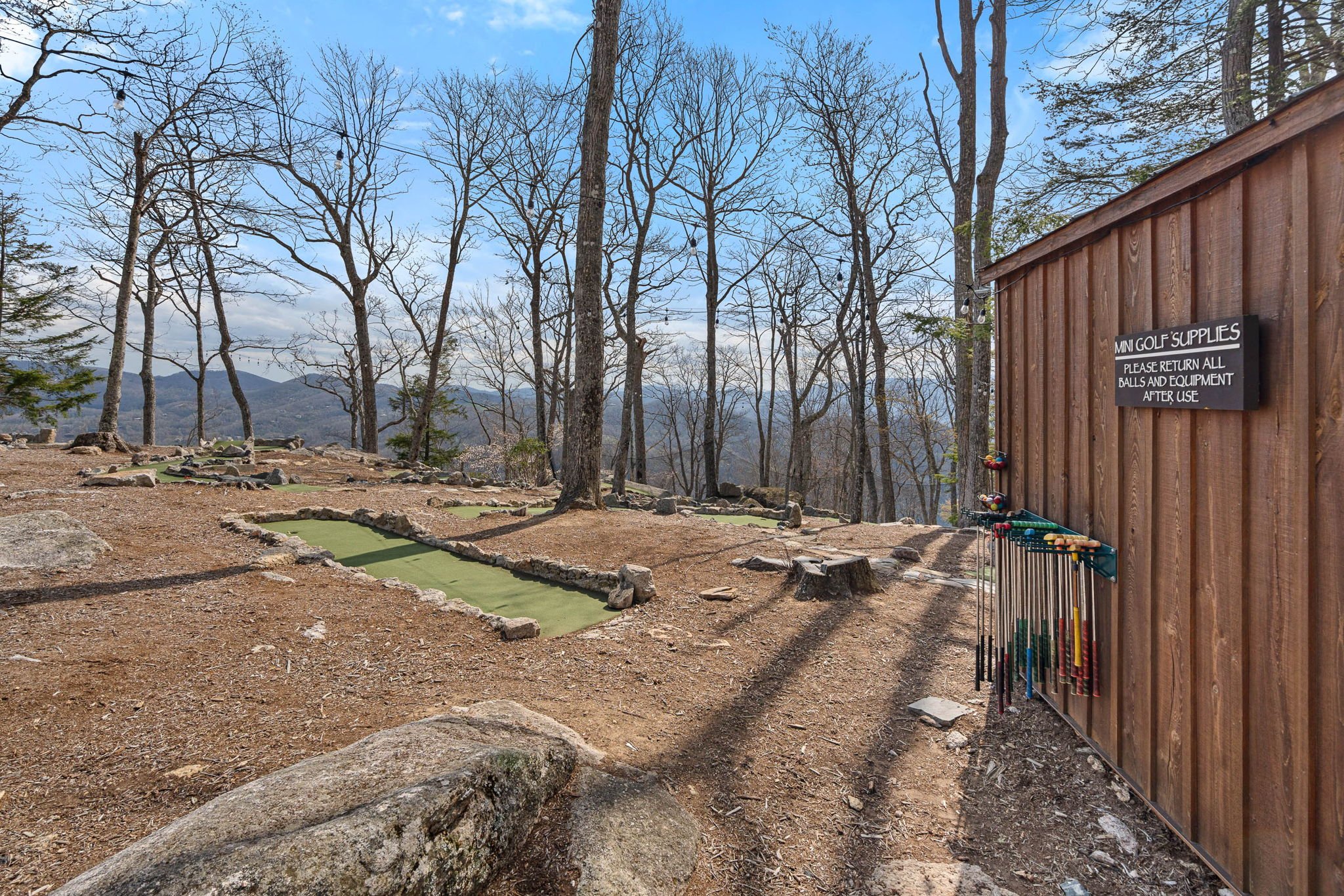 Outdoor mini golf course with a wooden shed that has a sign advising to return balls and equipment, leafless trees in the background, and a clear sky with clouds.