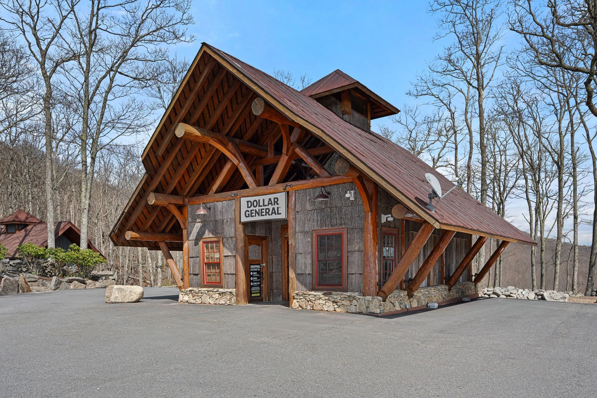 A wooden building with a sign that reads 'Dollar General' is situated in a rural wooded area with leafless trees and a clear blue sky.