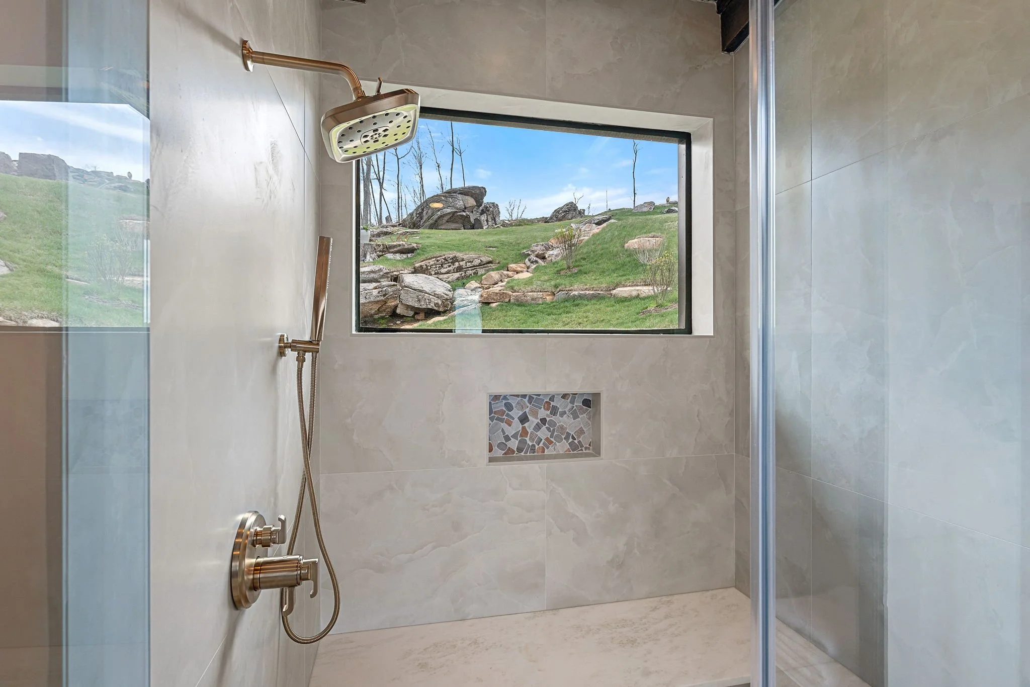 Shower area with a view of a rocky hillside landscape through a large window, featuring a rainfall showerhead and beige tiled walls with a decorative tile inset.