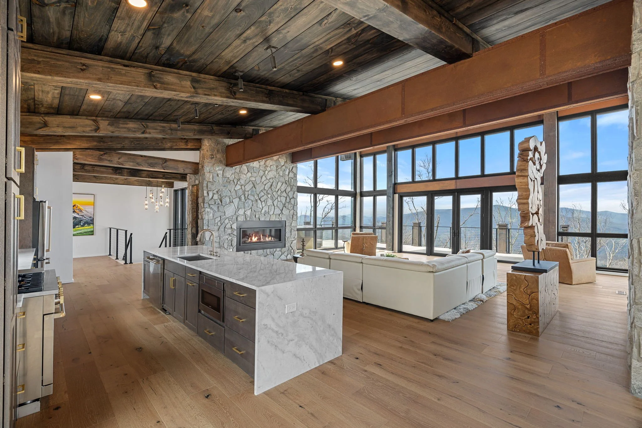 Modern open-concept living space with a kitchen island, white sectional sofa, large windows with mountain views, wood ceiling, and stone fireplace.