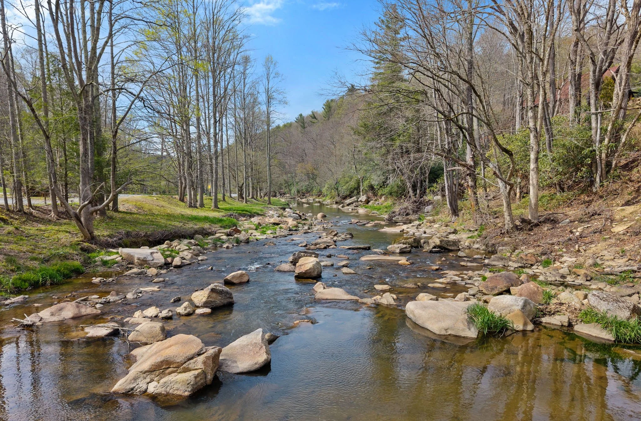 A creek flowing through a wooded area with trees on both sides, some with leaves and some without, under a blue sky with a few clouds.