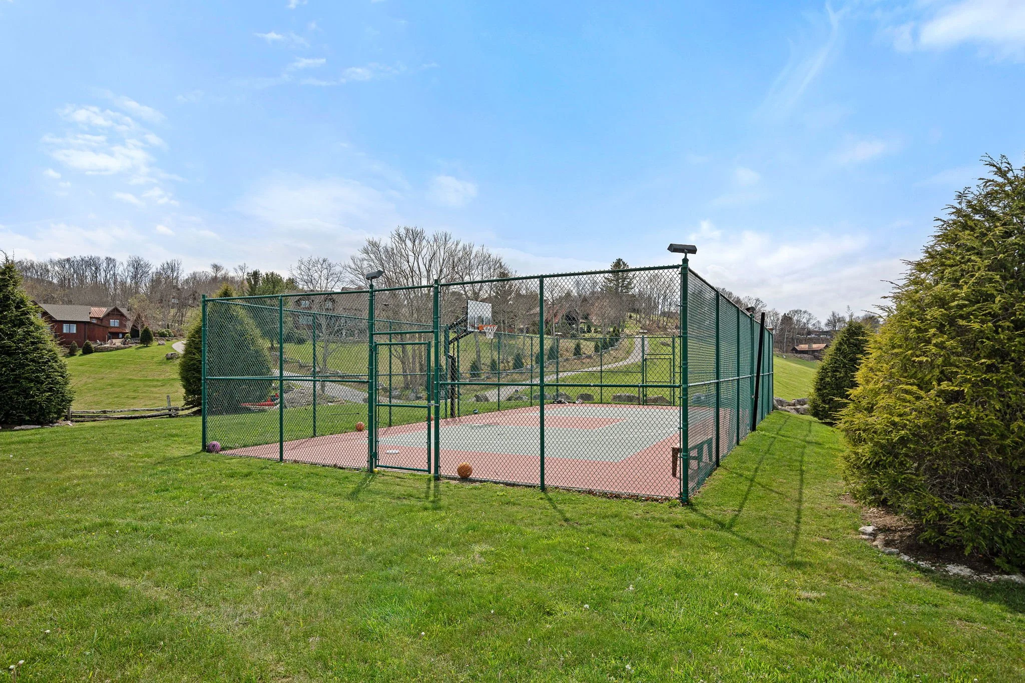 An outdoor basketball court surrounded by a green fence, with trees and houses in the background and a clear blue sky above.
