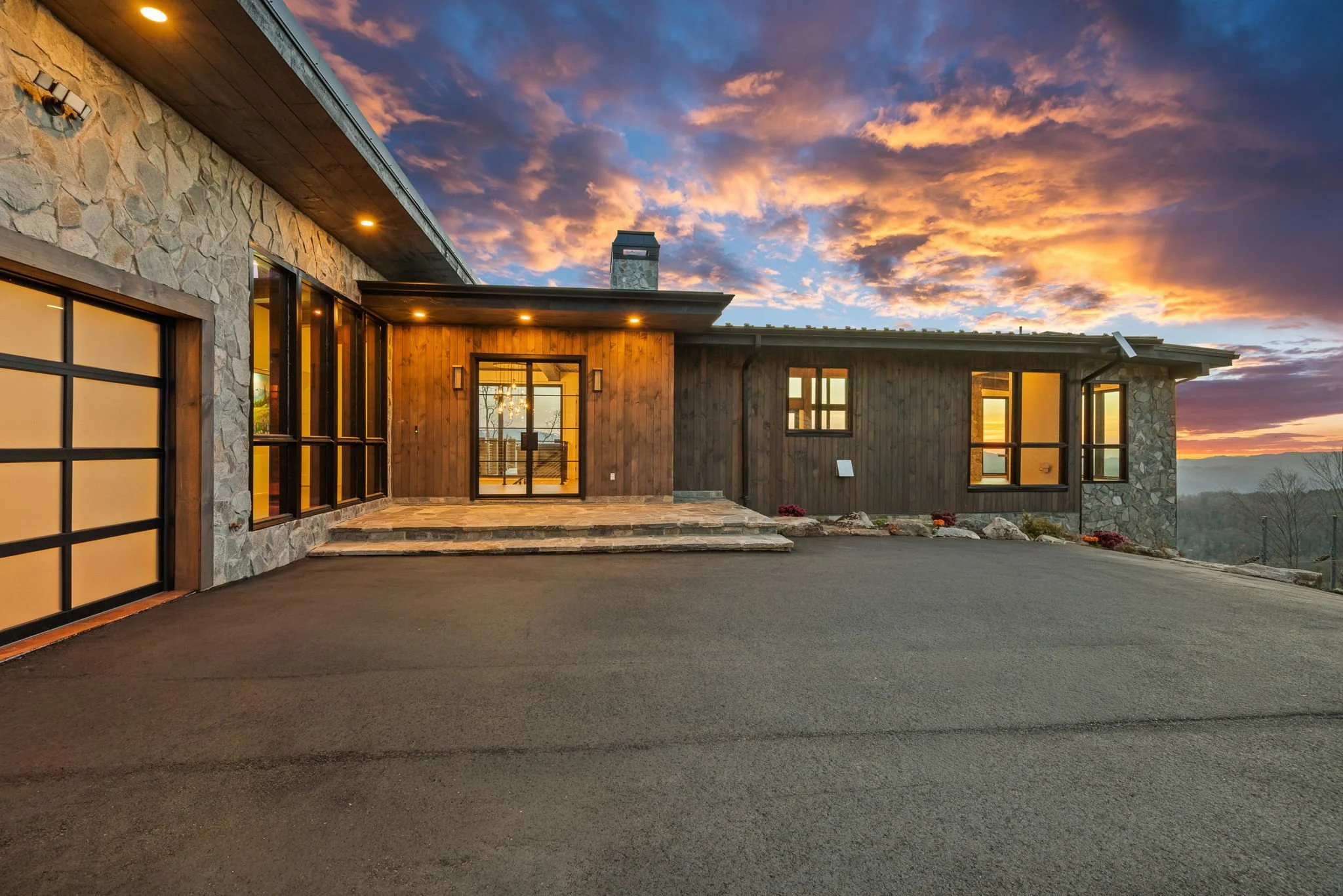 Exterior view of a modern house at sunset, with stone and wood siding, large windows, and a paved driveway.