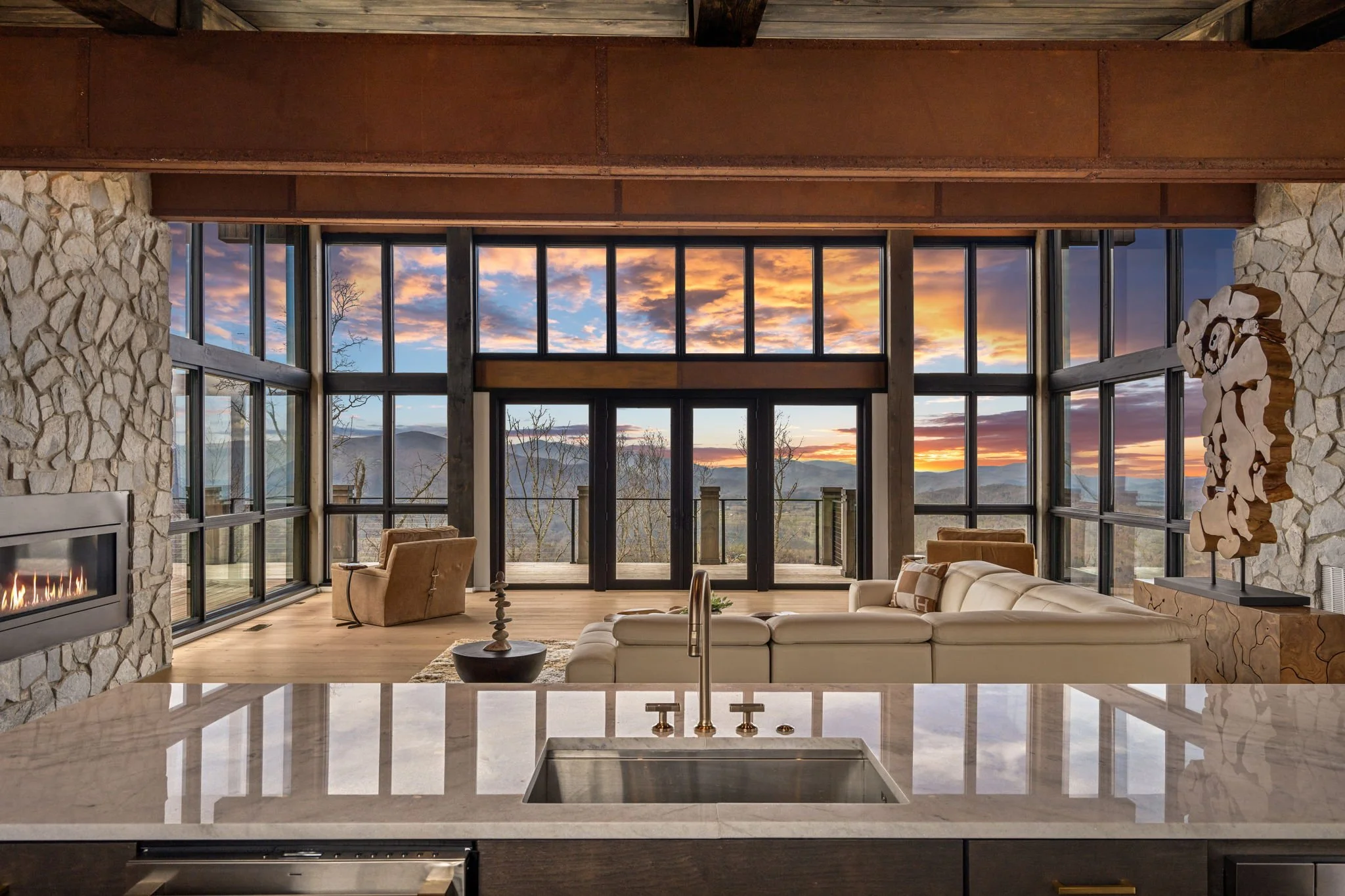 Living room with stone walls, large windows showing a mountain landscape during sunset, a white sofa, an armchair, a coffee table, and a modern fireplace.