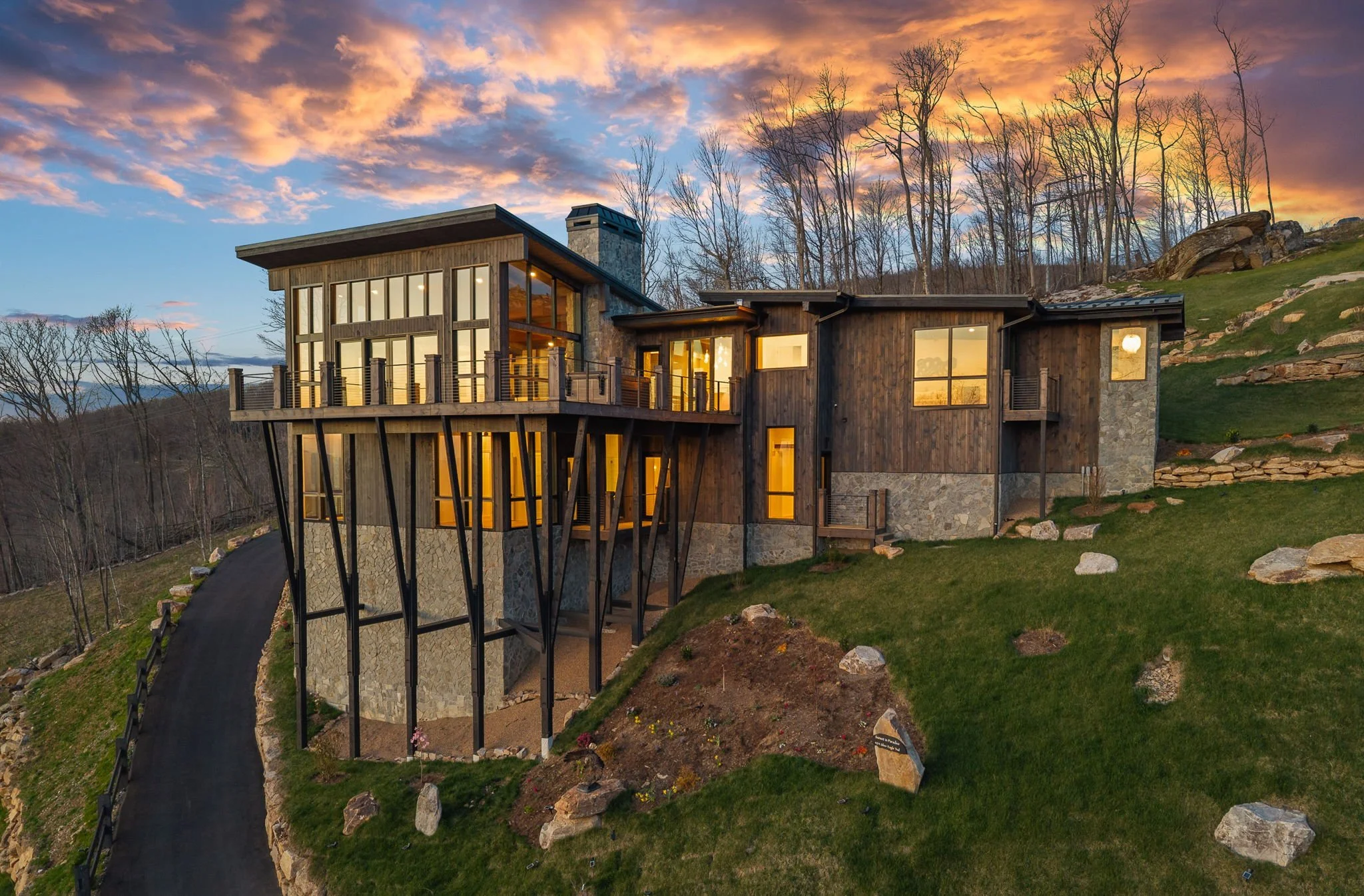 Modern house on a hillside at sunset, with large glass windows, wooden exterior, a chimney, scenic wooded landscape, and a driveway leading up to the house.