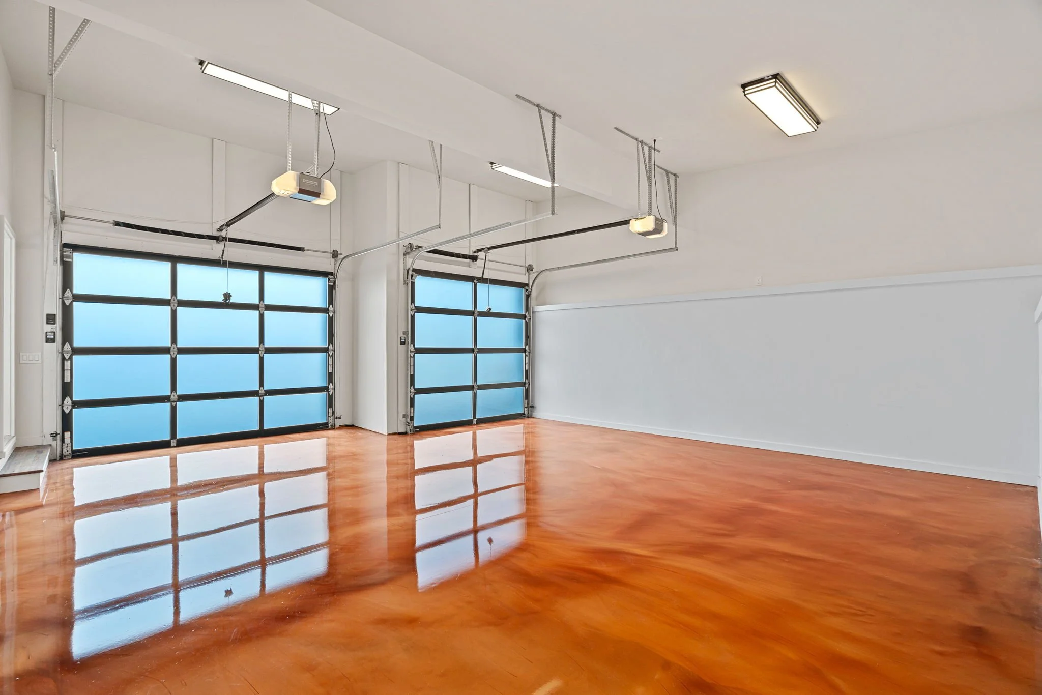 Empty garage with polished wooden floor, two blue-tinted glass garage doors, and ceiling-mounted garage door openers.