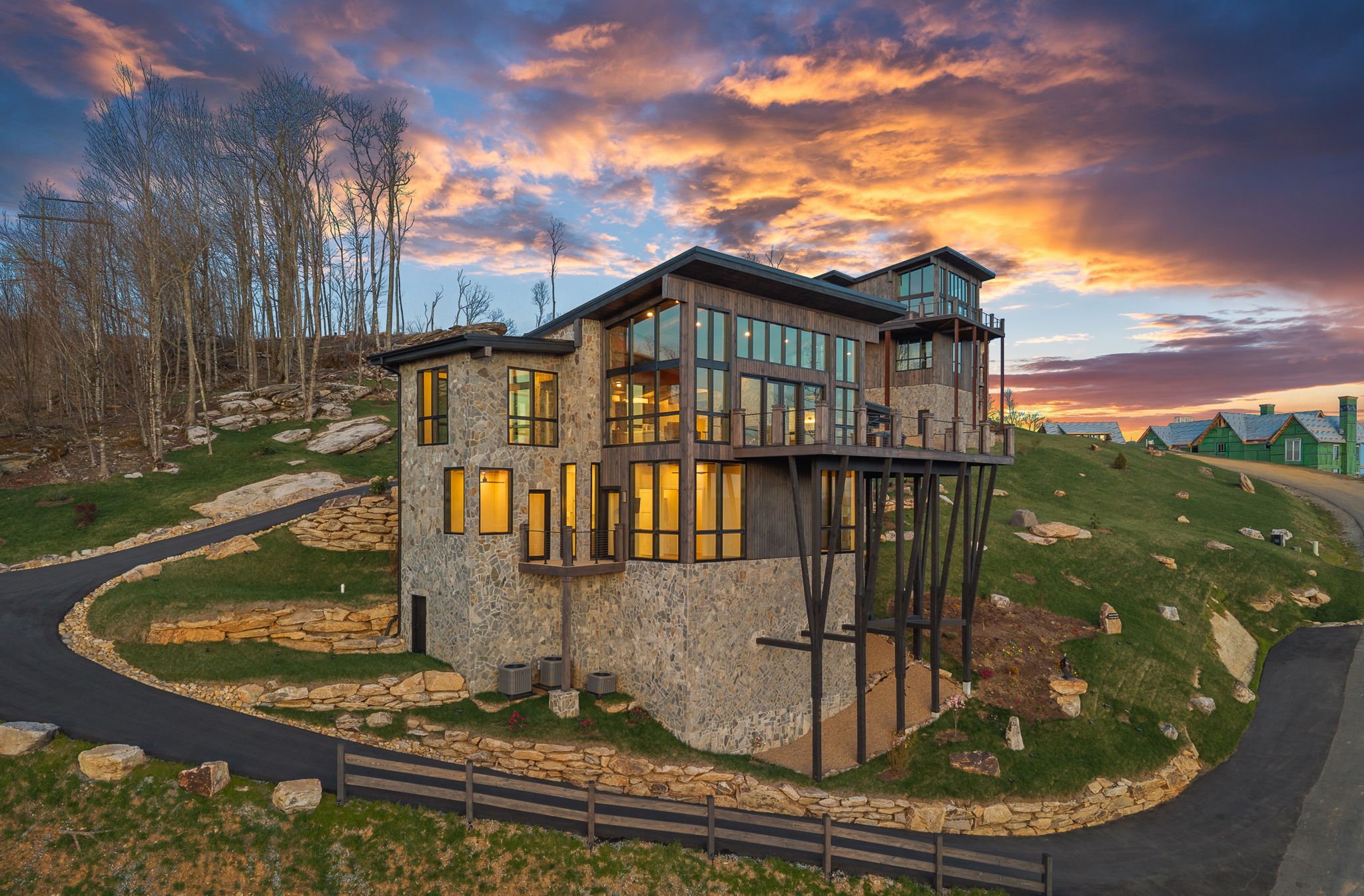 Modern house with large glass windows and stone exterior situated on a hill at sunset, with a winding driveway and neighboring houses in the background.