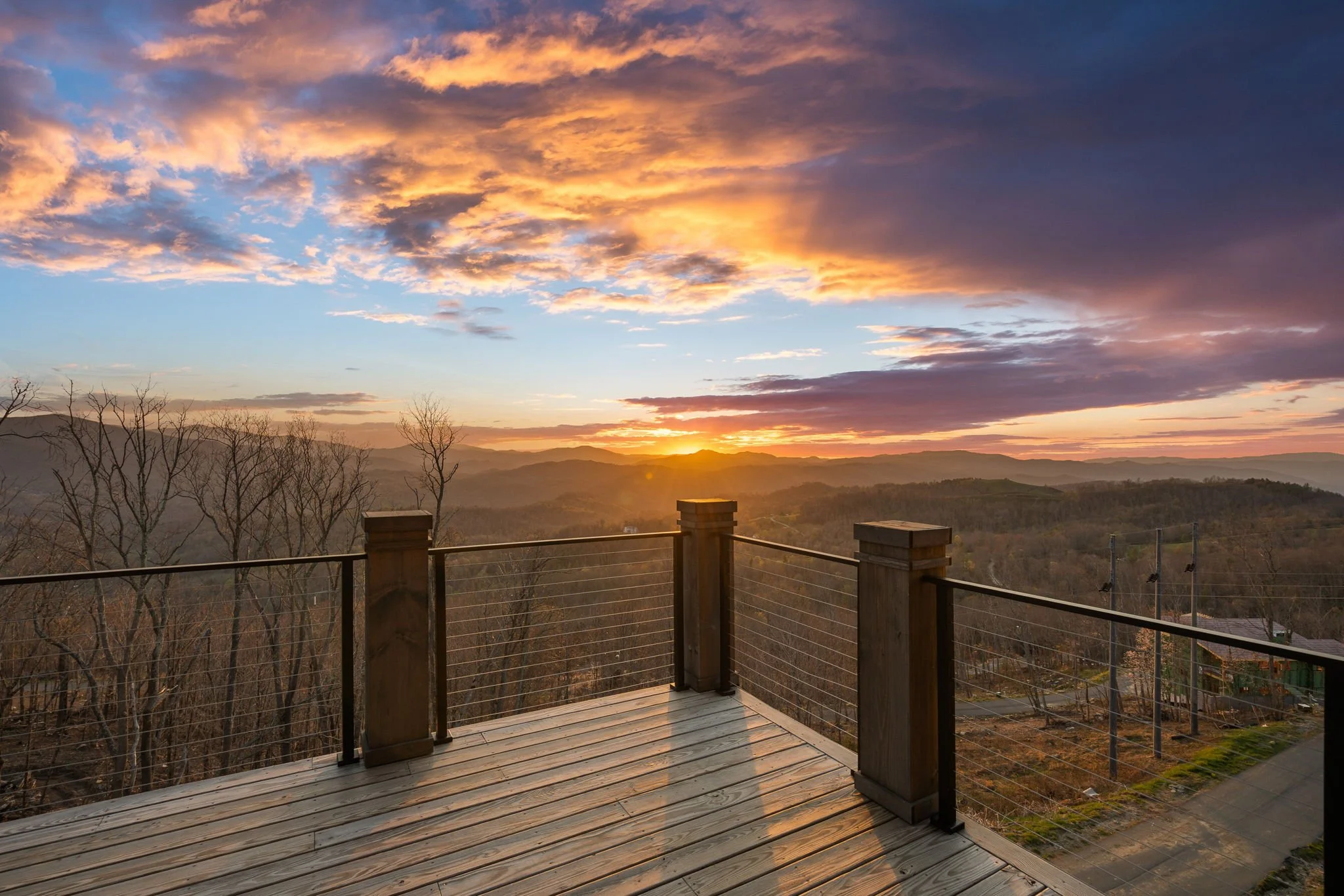 Sunset view from a wooden deck with a railing, overlooking hilly terrain and a sky filled with colorful clouds.