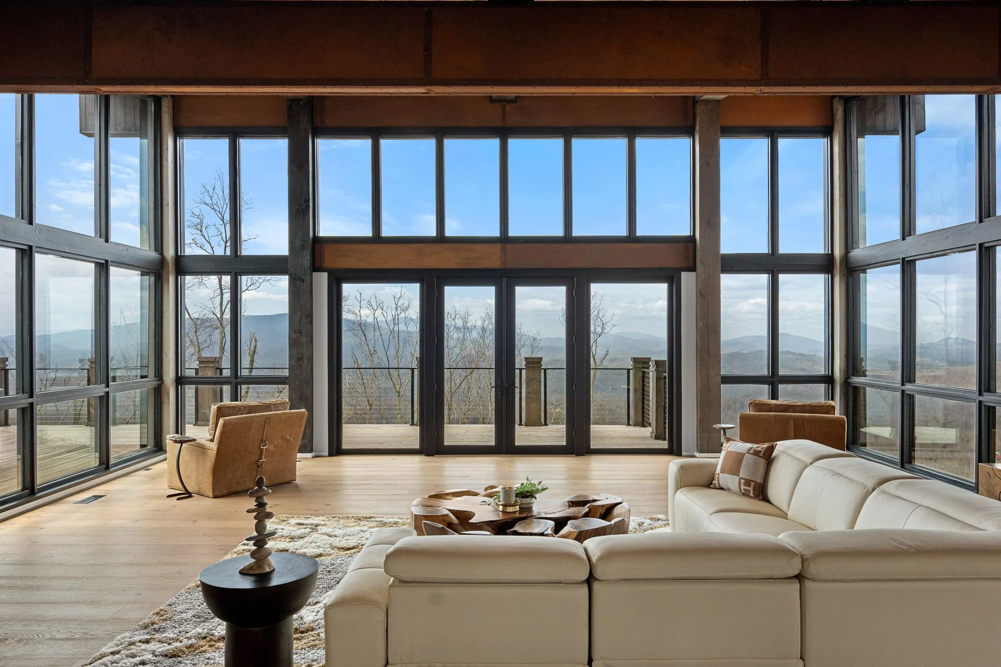 Living room with large floor-to-ceiling windows overlooking a mountain landscape, modern beige sectional sofa, wooden coffee table, and armchair.