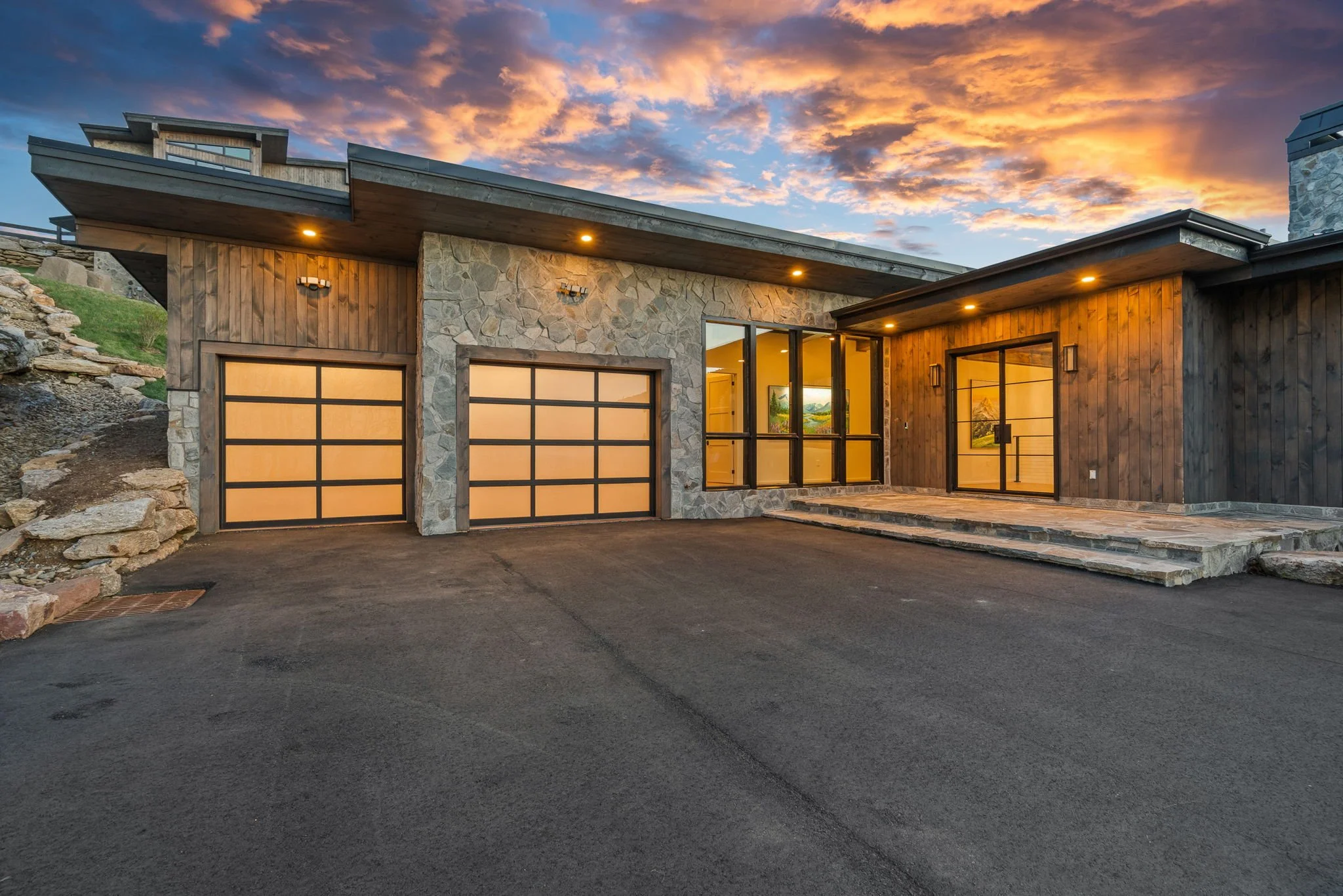 Modern house with stone and wood exterior, large glass doors, driveway, and sunset sky.