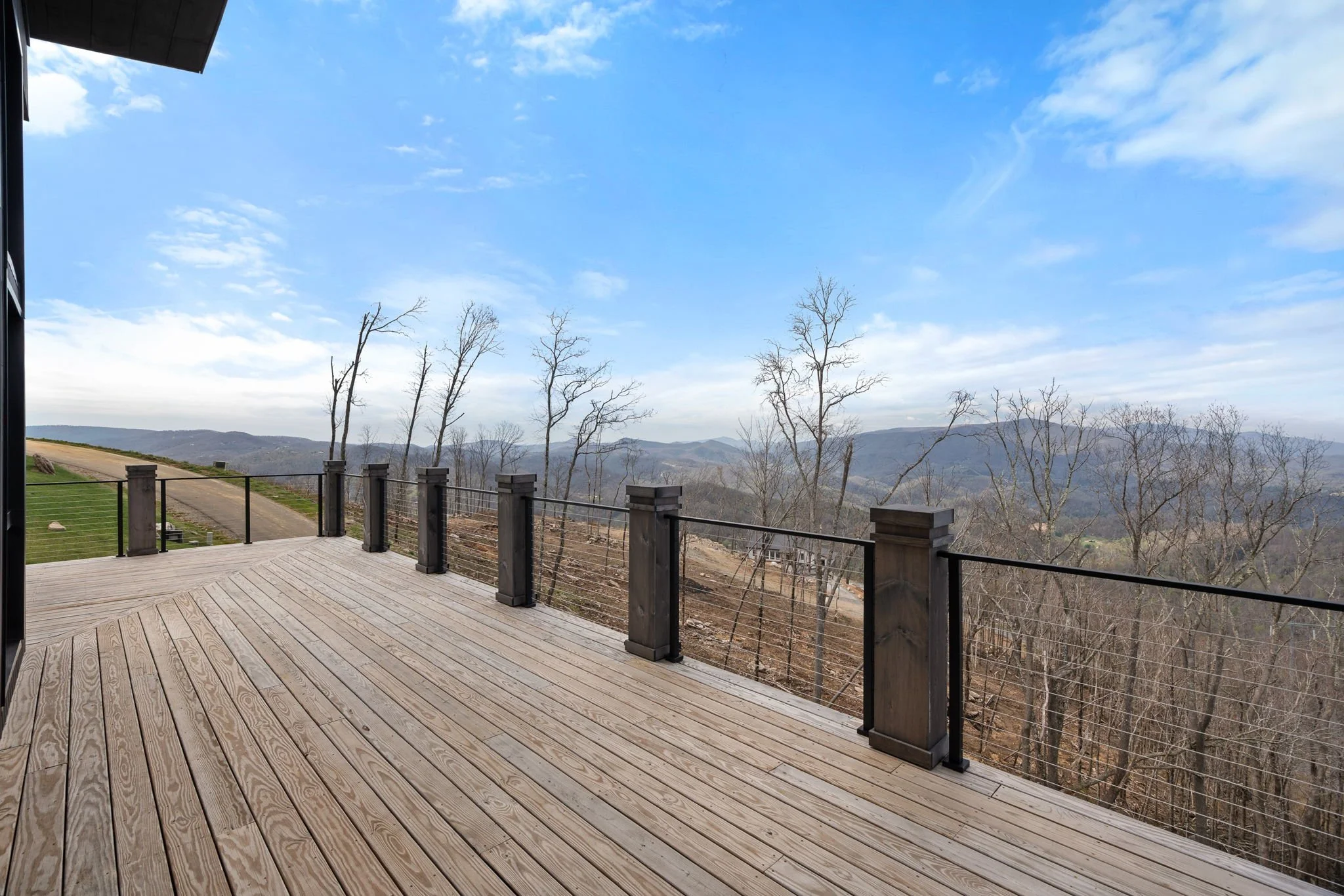 Wooden deck with black metal railing overlooking leafless trees and distant mountains under a partly cloudy blue sky.