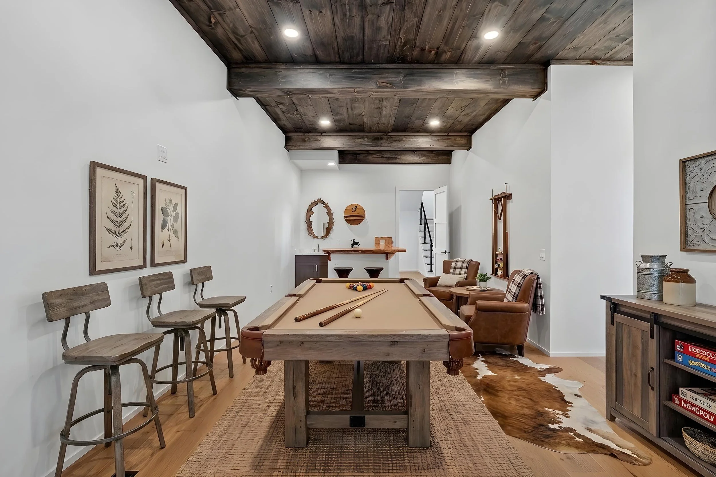 A basement room with a pool table, wood flooring, and white walls decorated with botanical artwork. There are four chairs along one wall, a cowhide rug, and a small bar area in the corner. The ceiling features wooden beams and recessed lighting.
