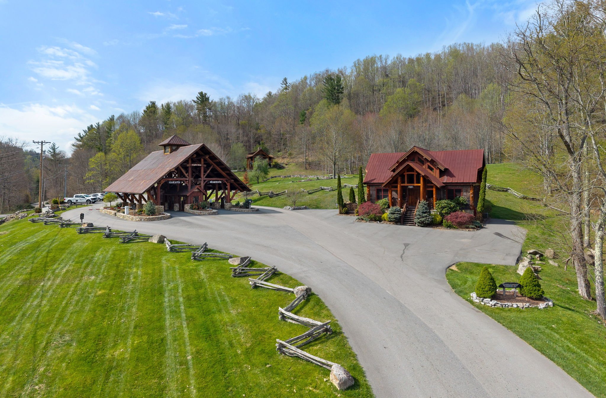 A rural lodge with a driveway, surrounded by a green lawn, trees, and a wooded hillside in the background.