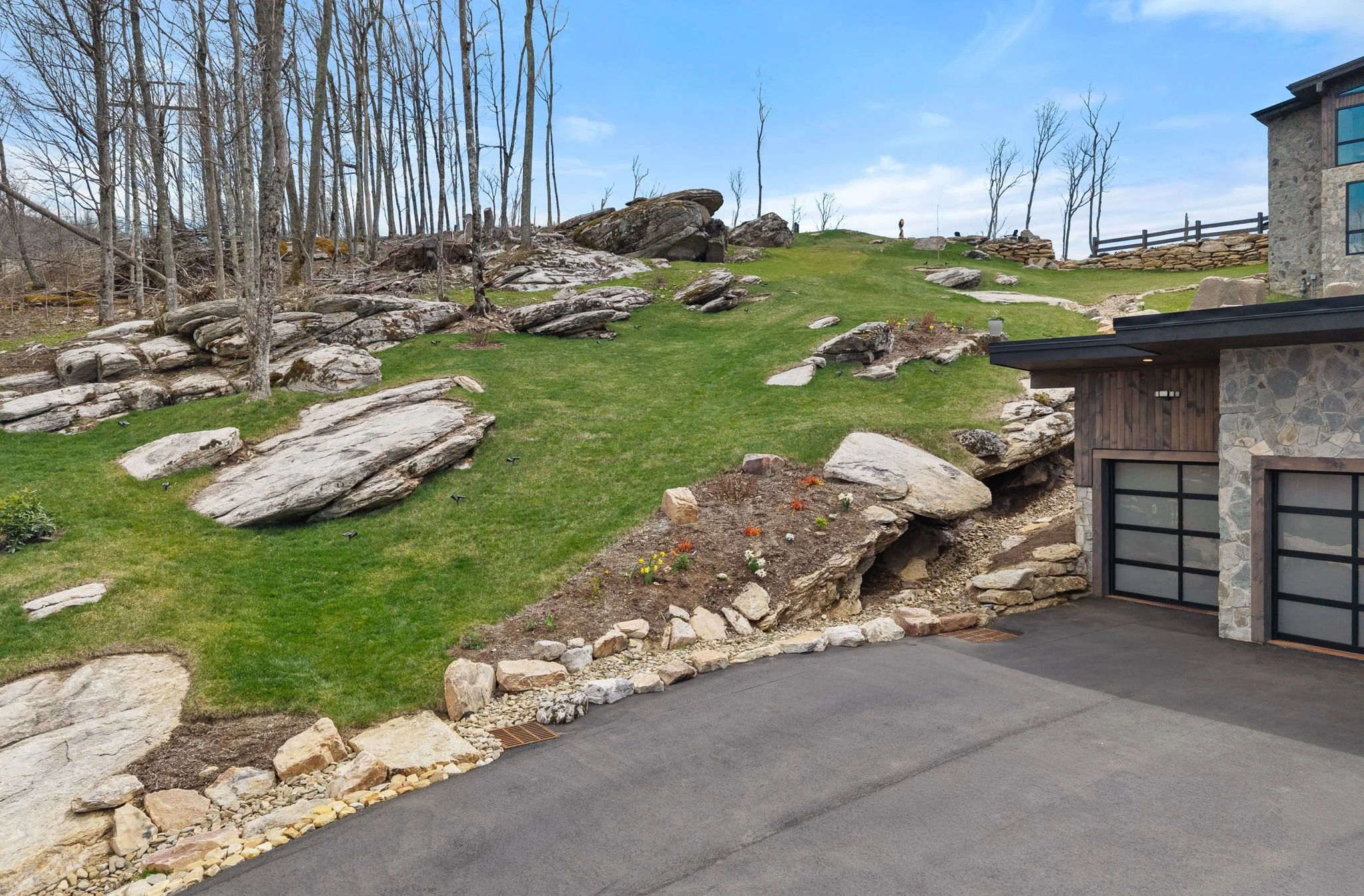 A residential backyard with a sloped grassy hill, large rocks, and trees. Part of a house with stone and wood exterior is visible in the foreground.