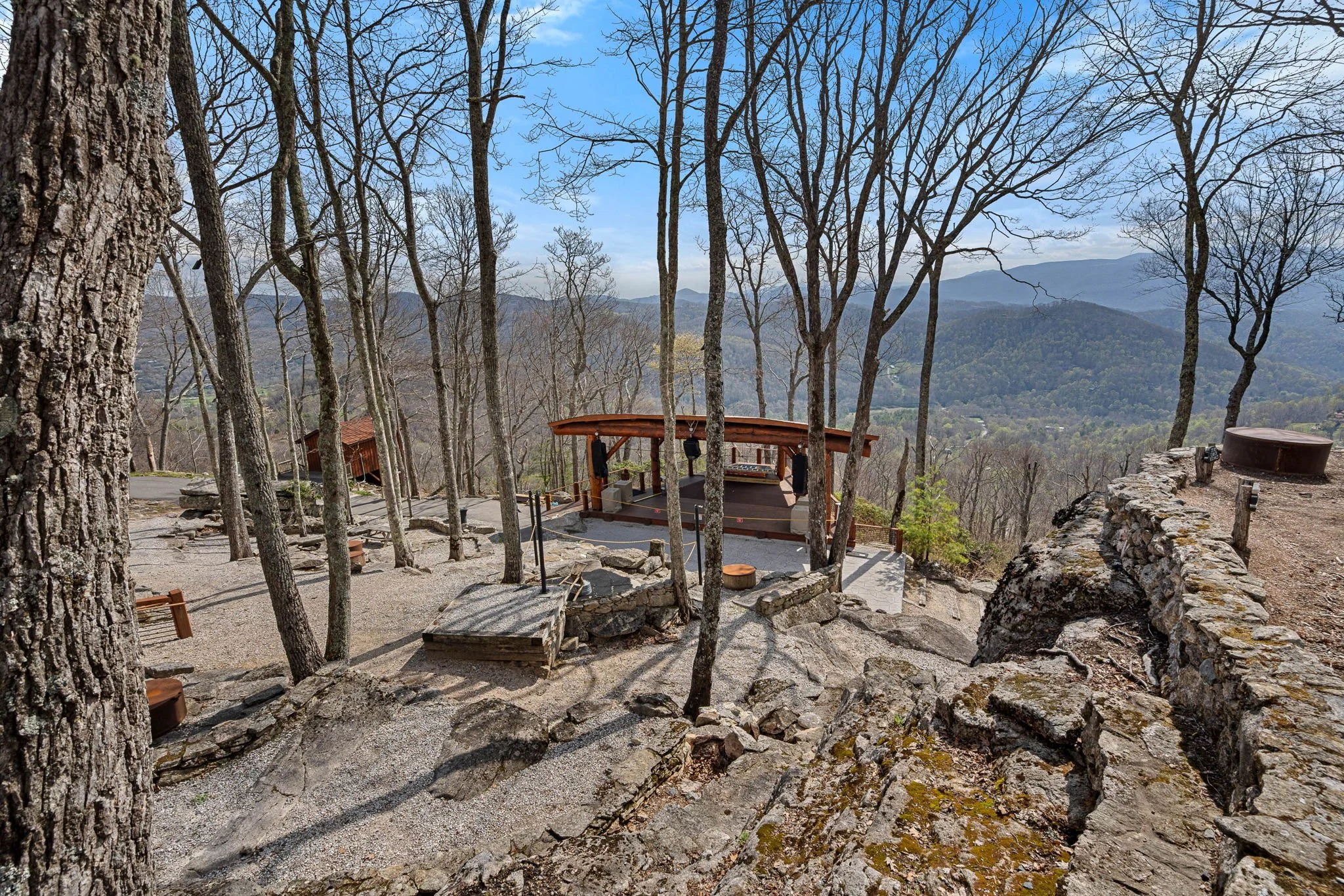 A scenic mountain overlook with leafless trees, a small wooden stage with audio equipment, and a panoramic view of distant mountains and a valley under a partly cloudy sky.