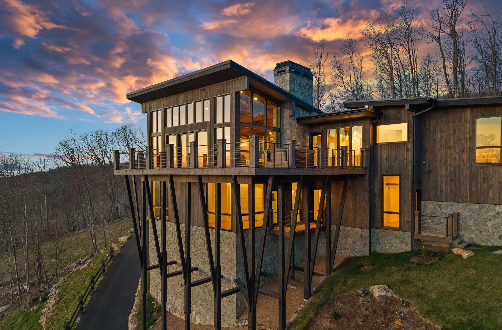 A modern house on a hillside during sunset with large windows, a stone chimney, a wooden balcony, and surrounding leafless trees.