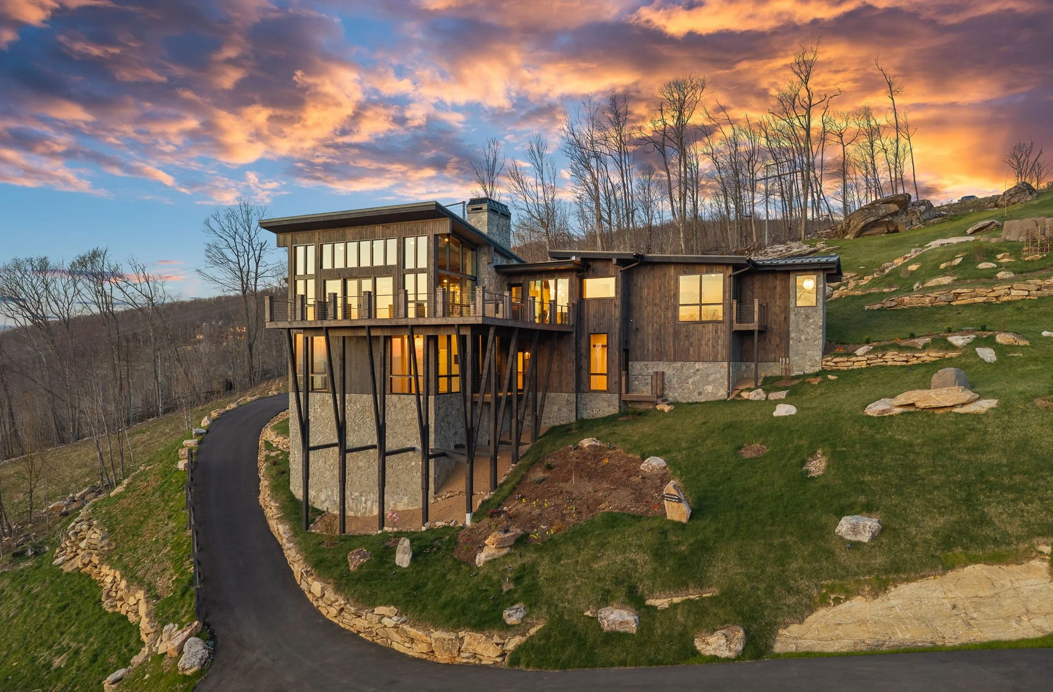 Modern house on a hillside with large windows, surrounded by a landscaped yard with rocks and a winding driveway, under a vibrant sunset sky.