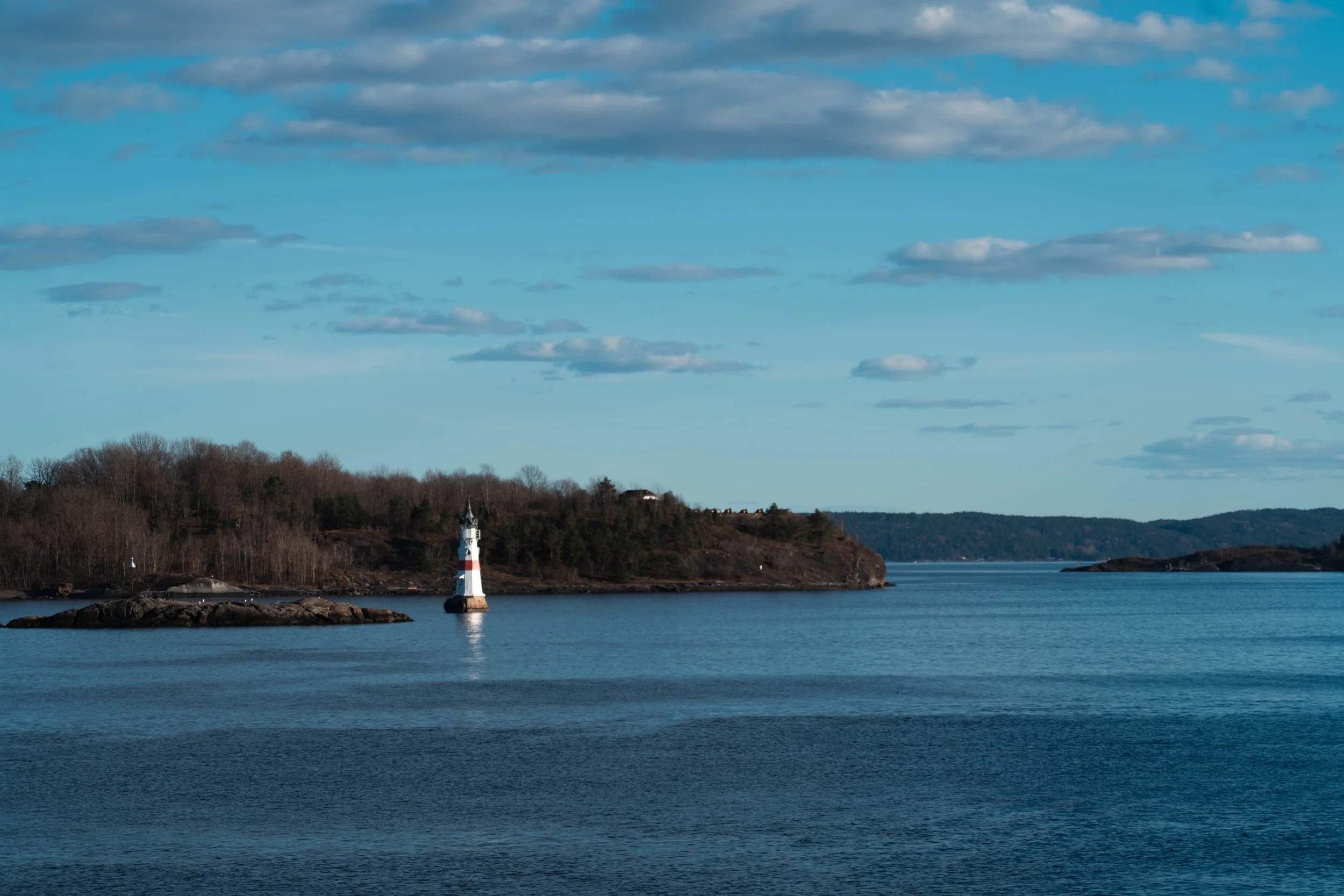 Scenic view of a lighthouse on a coastal hillside with trees, overlooking calm water under a blue sky with scattered clouds.