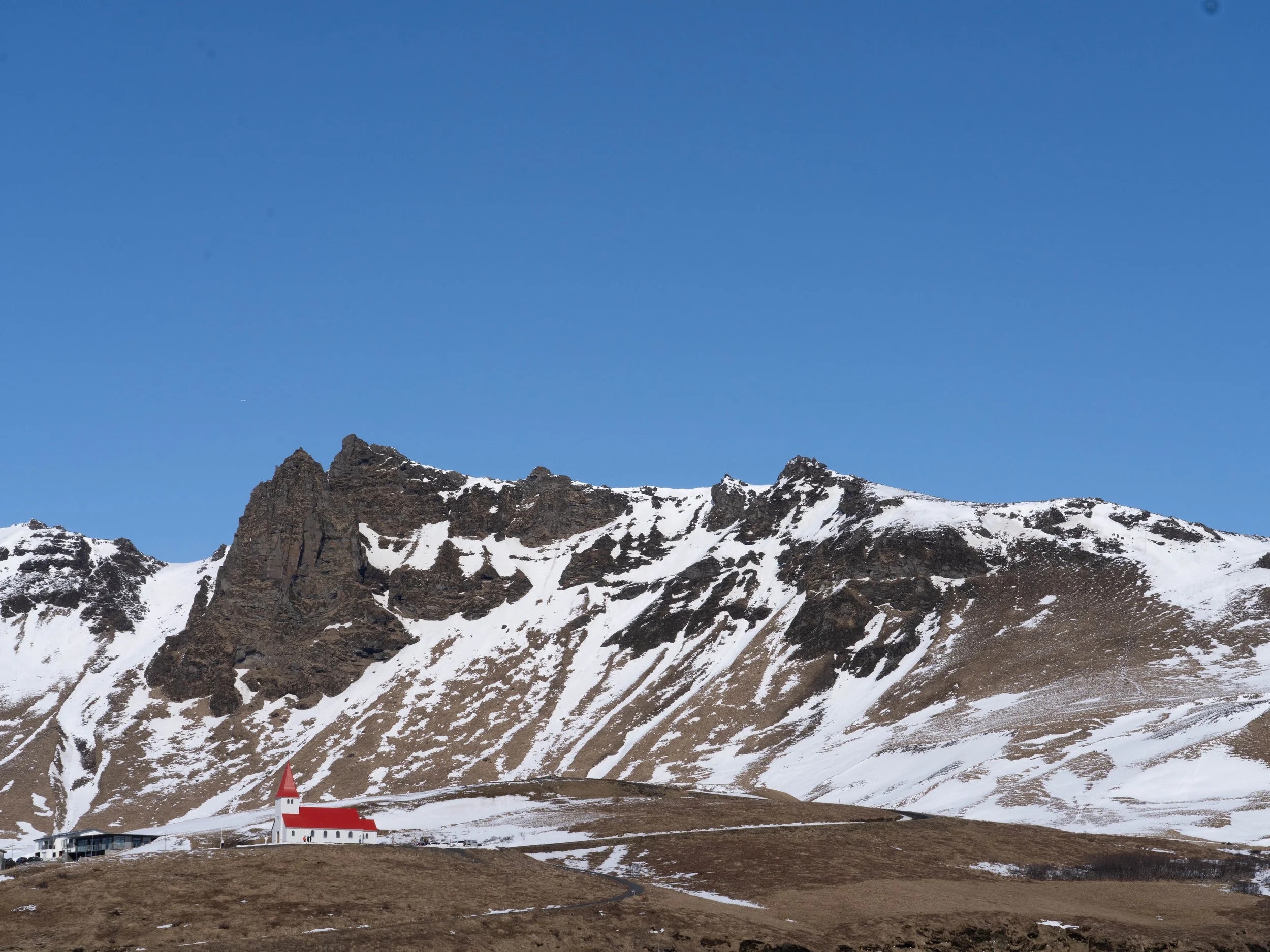 Snow-covered mountain with a small white church featuring a red steeple and roof in a valley at the base of tall jagged rocky mountains with patches of snow under a clear blue sky.