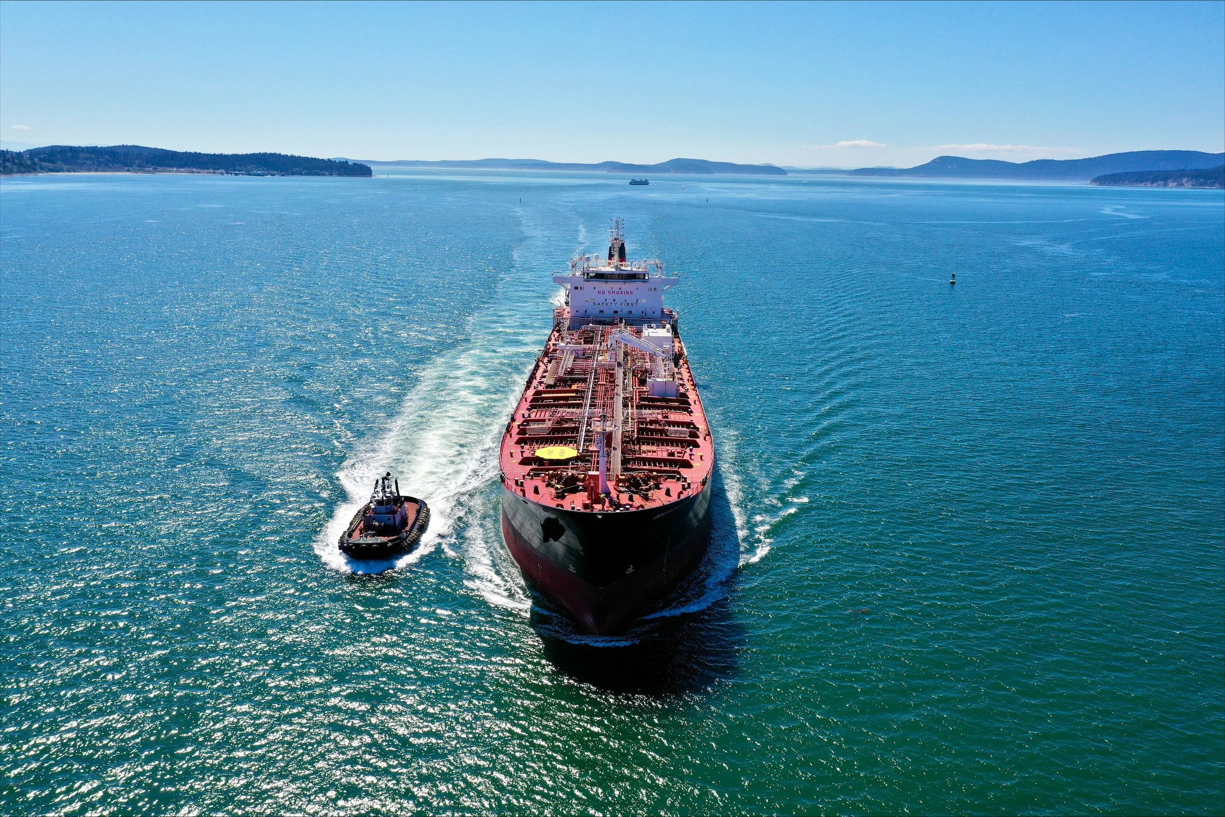 A large oil tanker ship being guided by two tugboats on a clear blue body of water with distant land and hills on the horizon under a clear sky.
