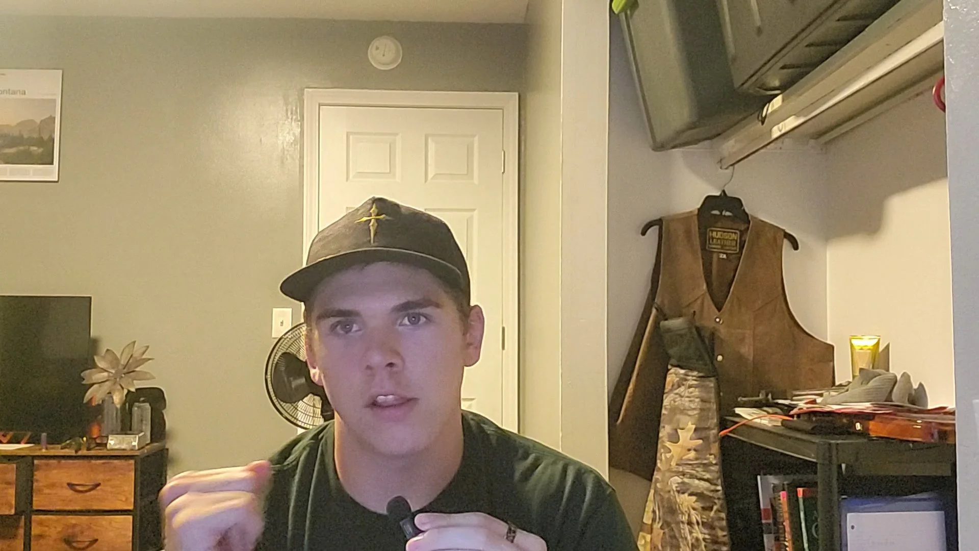 Young man wearing a black baseball cap with a yellow cross and a camouflage shirt, sitting indoors with a background of a dresser, a fan, a wall with a poster, and various objects.