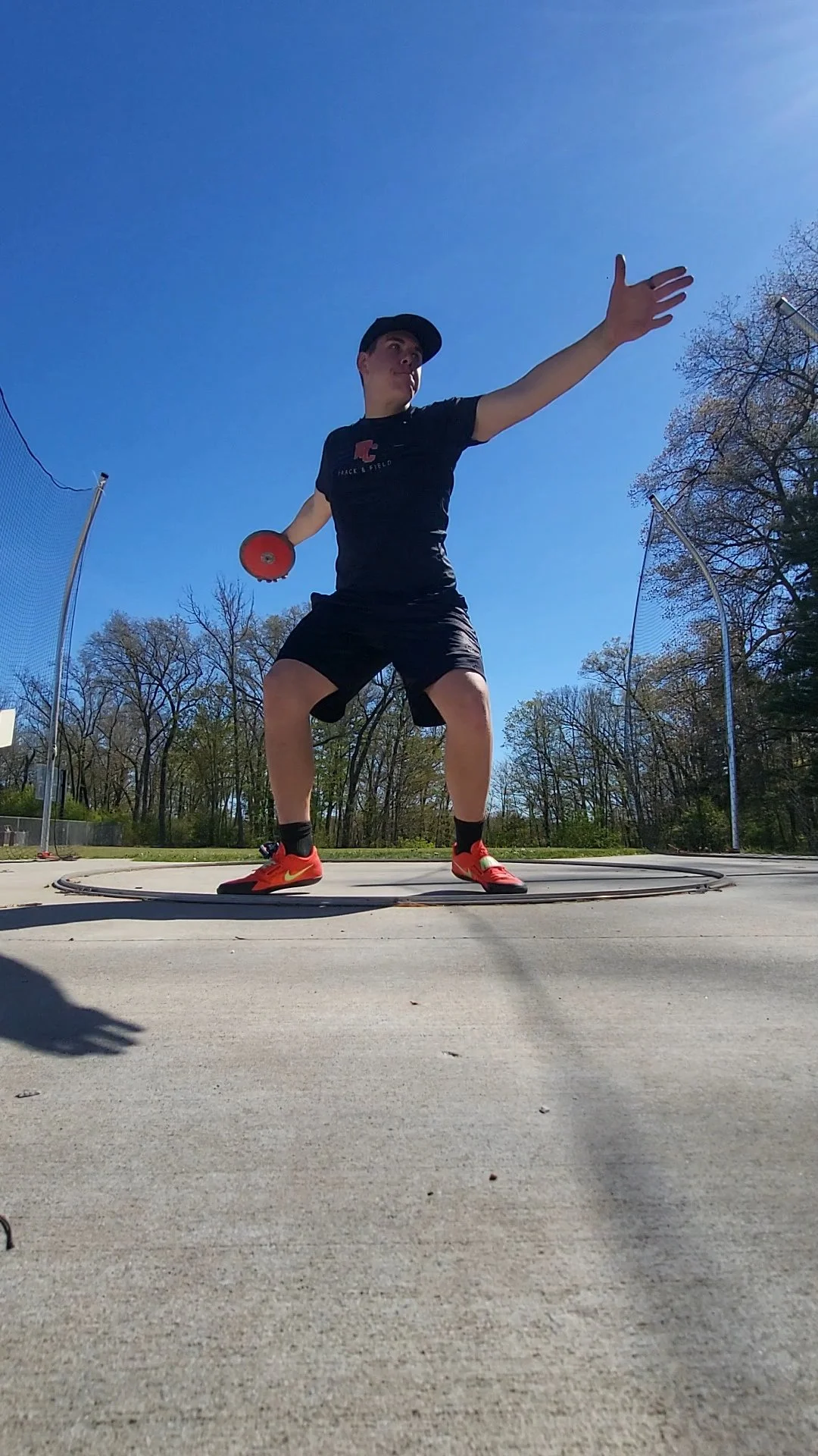 A young man dressed in black is practicing discus throw outdoors on a sunny day, standing in a discus circle with trees and a blue sky in the background.