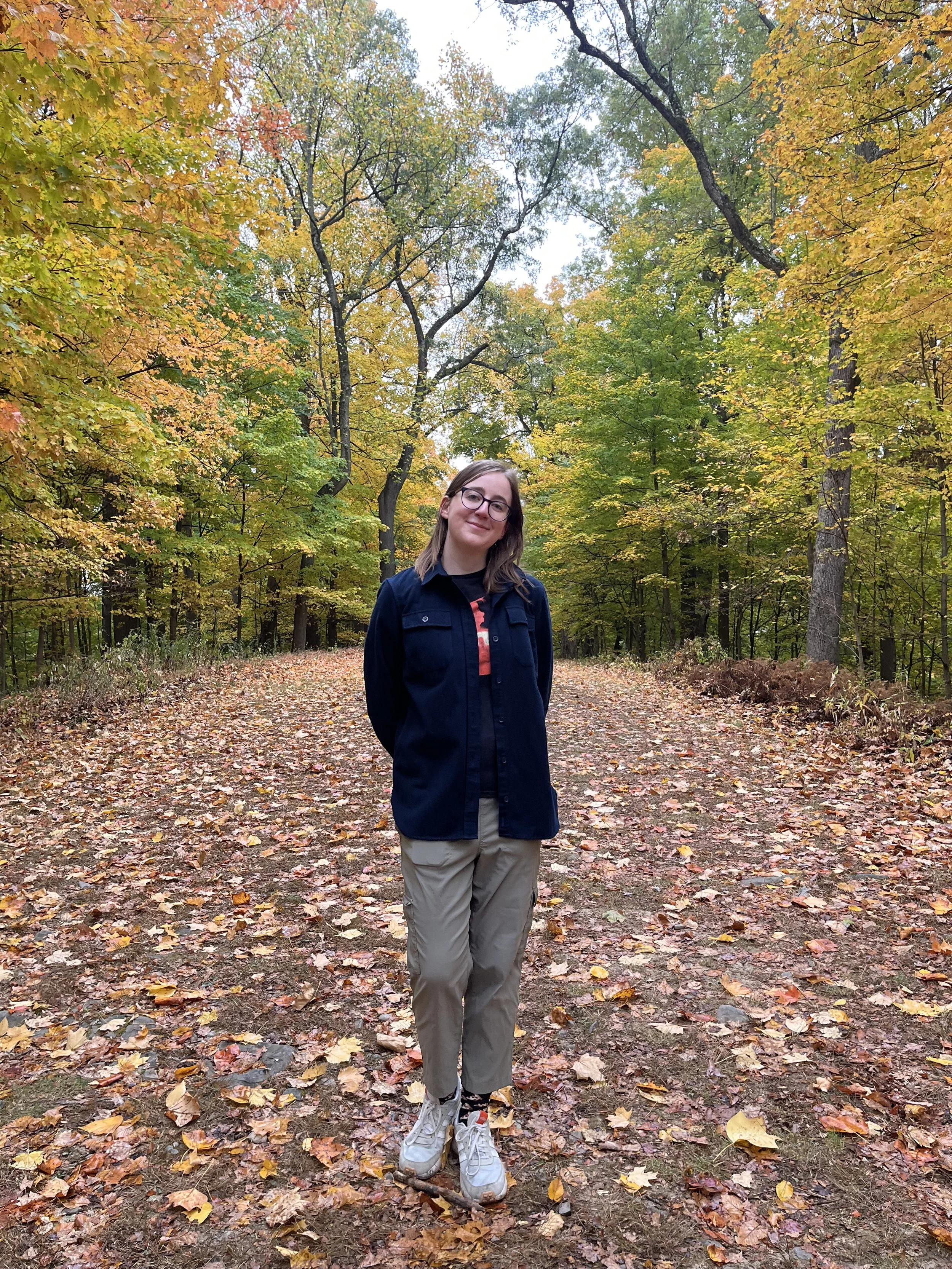 A person standing on a path covered with fallen autumn leaves in a forest with trees showing green and yellow foliage.