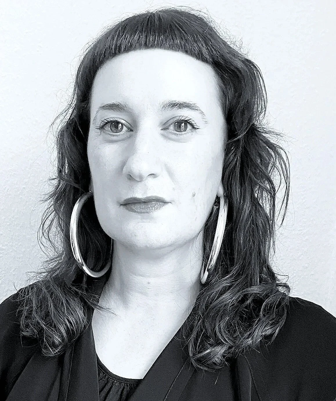A black and white portrait of a woman with wavy hair, wearing large hoop earrings and a dark top, against a plain background.
