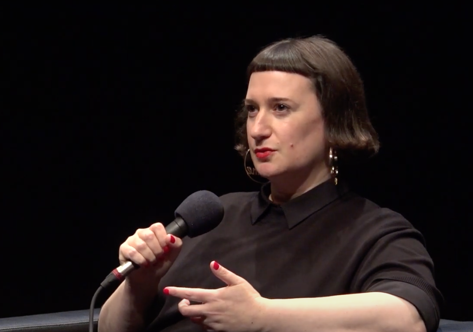 A woman with short dark hair, wearing a black top and hoop earrings, holding a microphone and speaking during an interview or a discussion.