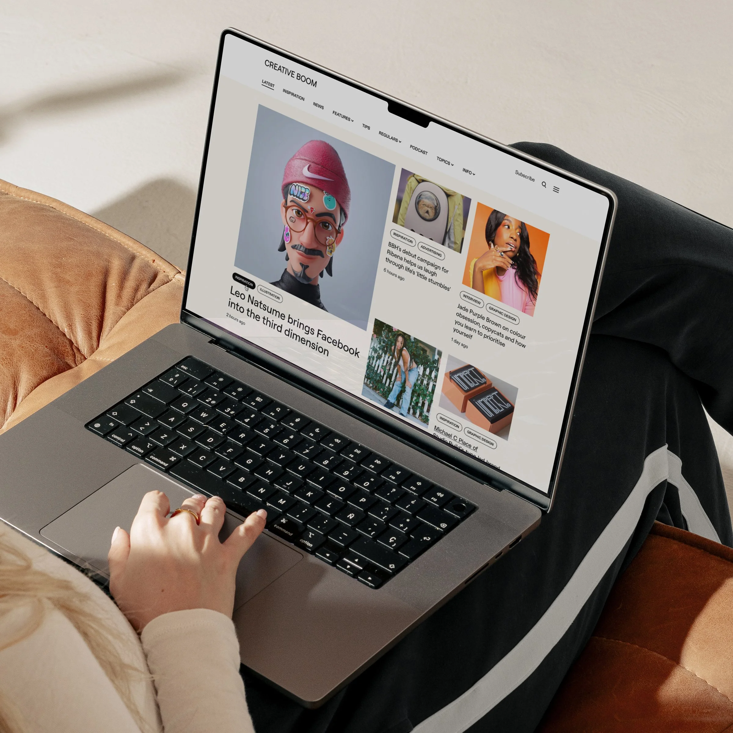 Close-up of a person's hand typing on a laptop keyboard, showing Creative Boom on the screen, with a magazine on the table and a beige couch nearby.