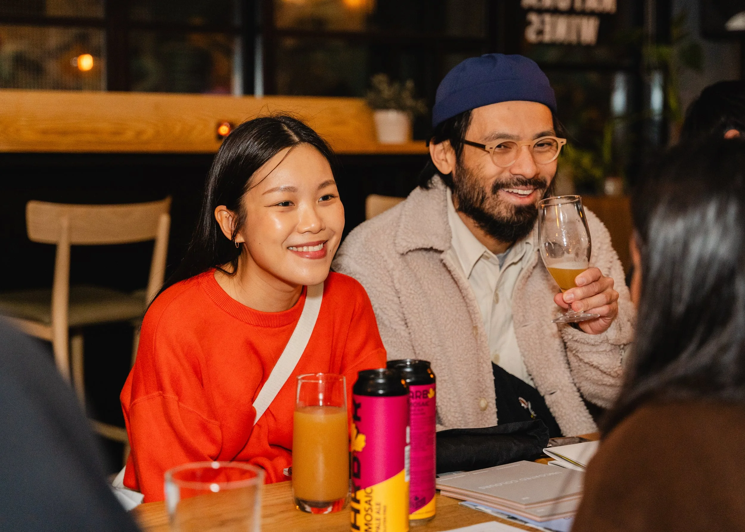 A group of people sitting at a table, smiling and enjoying drinks at a social gathering or dinner. A woman in an orange sweater and a man wearing glasses and a blue hat are visible.