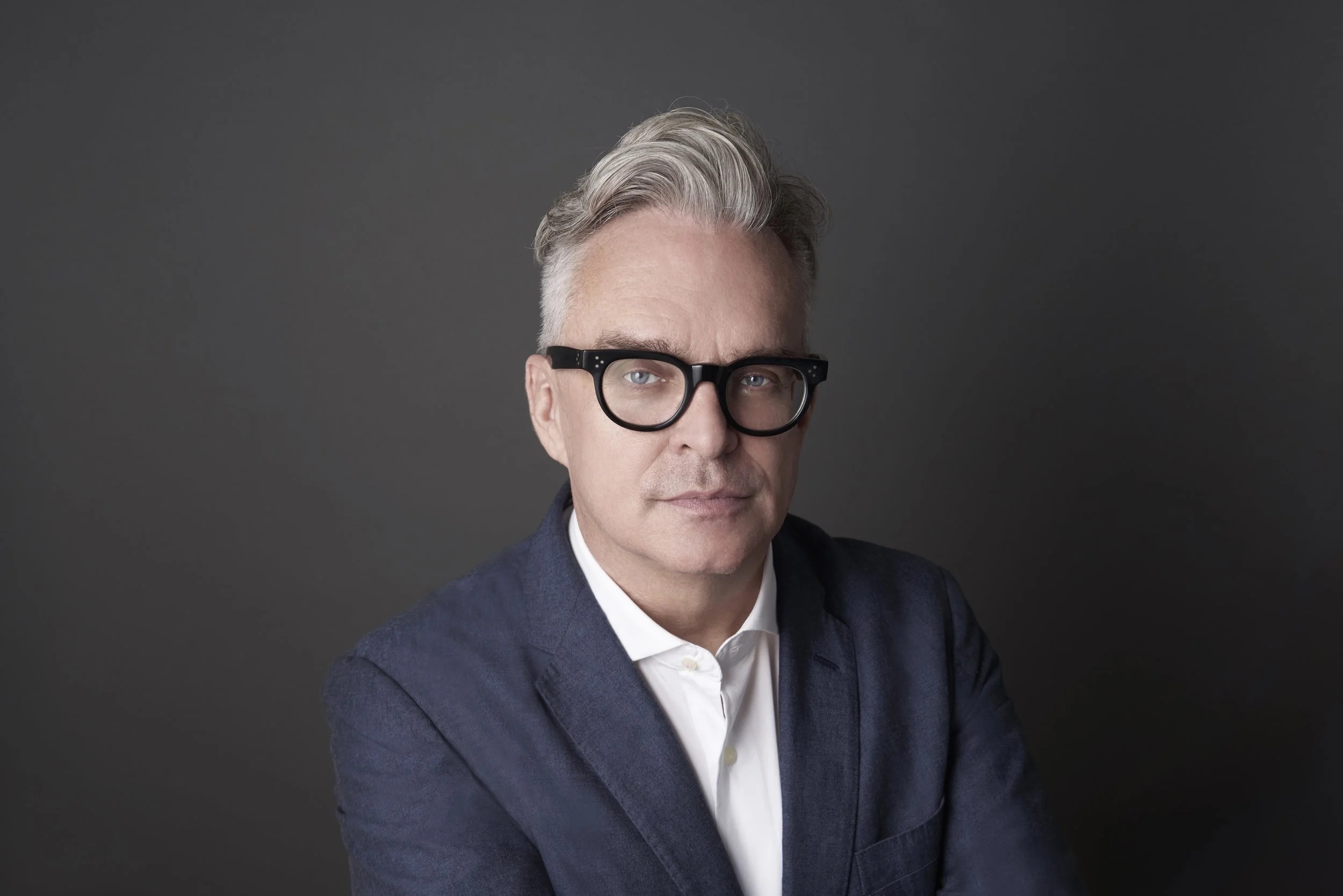 Portrait of Brian Collins, who has grey hair and black glasses, wearing a navy blazer and white shirt against a plain dark background.