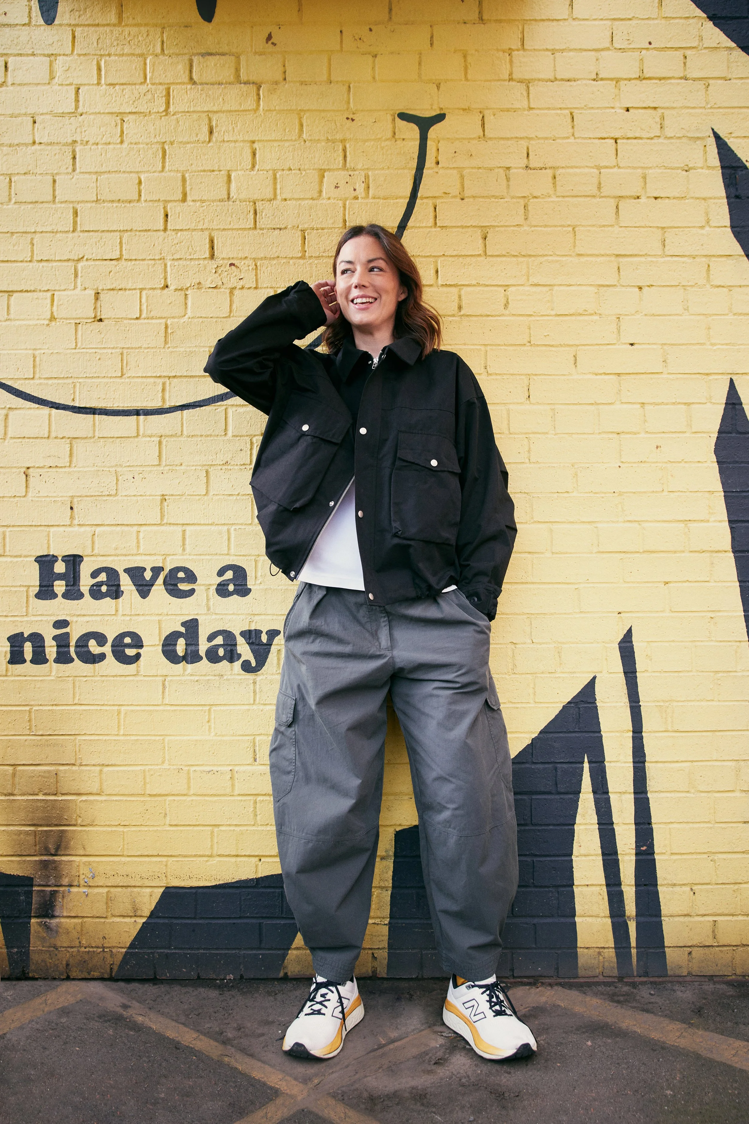Katy Cowan is standing in front of a yellow brick wall with black and white text, wearing a black jacket, grey cargo pants, and white sneakers with black and yellow accents, smiling and looking to her left.