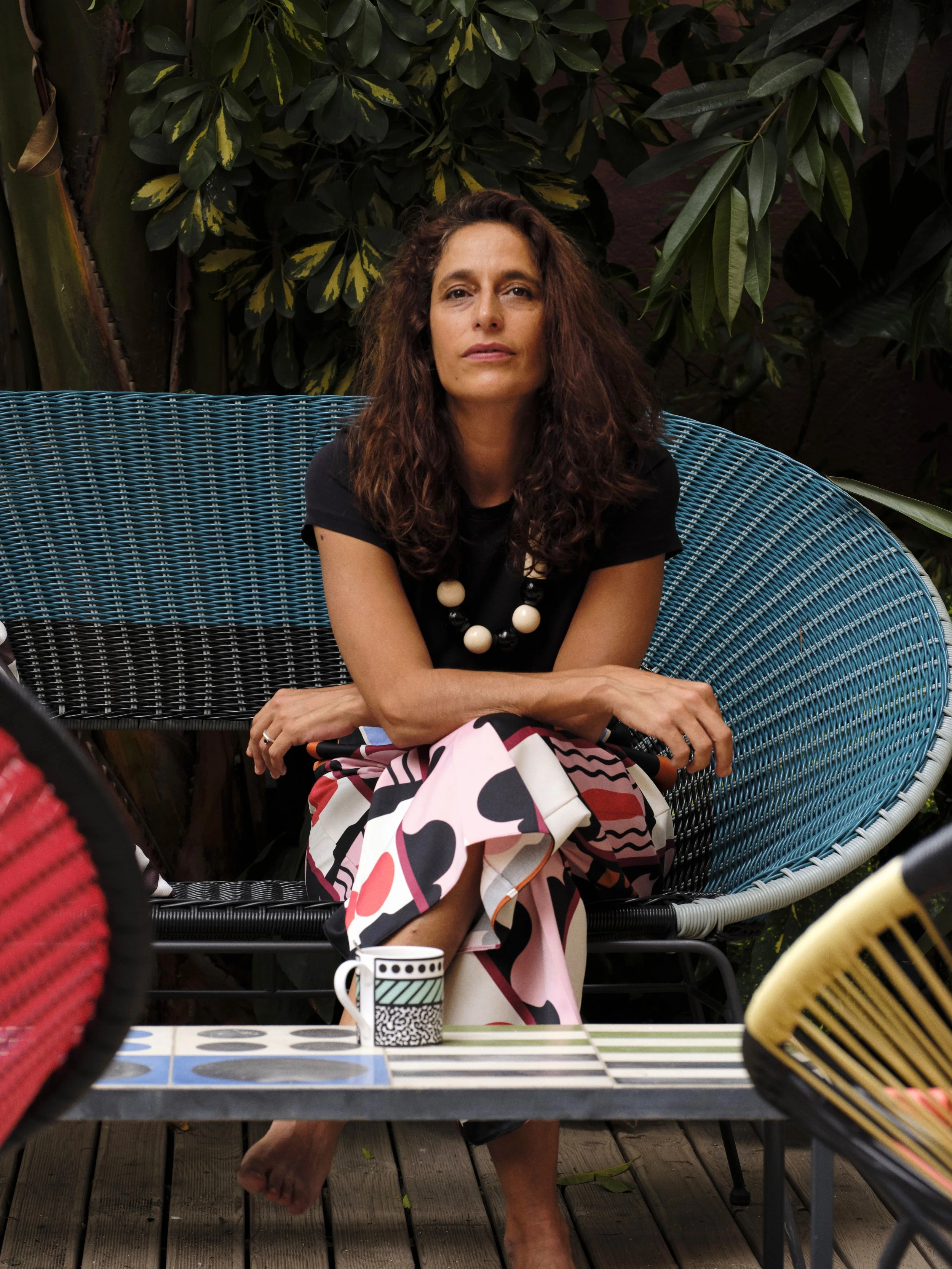 Malika Favre, sitting under lush green foliage, wearing a black top and a black and white beaded necklace, with colour-patterned pants. She sits on a blue woven outdoor chair.