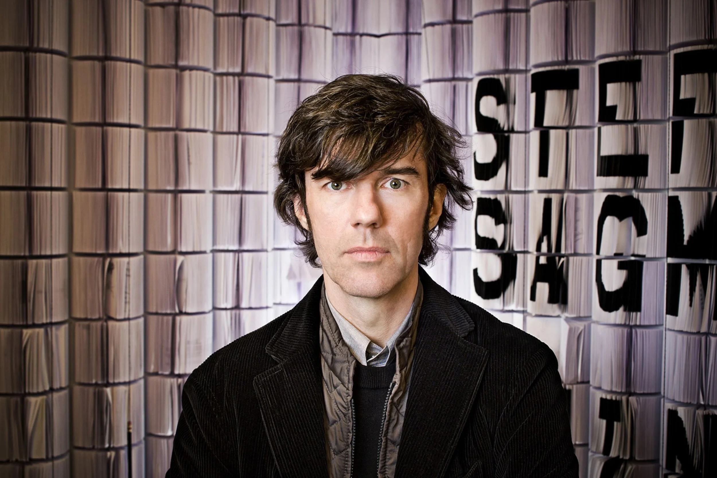 Stefan Sagmeister, a man with dark hair and a serious expression, is standing in front of a background of large stacked paper rolls with a partial sign that reads 'SEN' and 'T'.