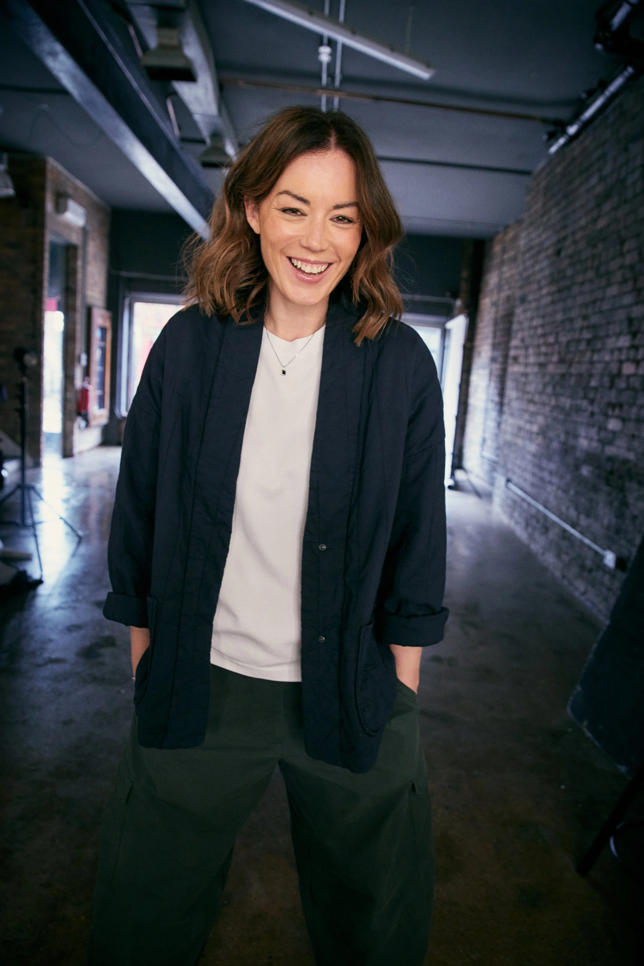Katy Cowan, a woman with shoulder-length brown wavy hair, wearing a dark jacket over a white shirt, standing in an industrial-style interior with exposed brick walls and hanging pipes, smiling at the camera.