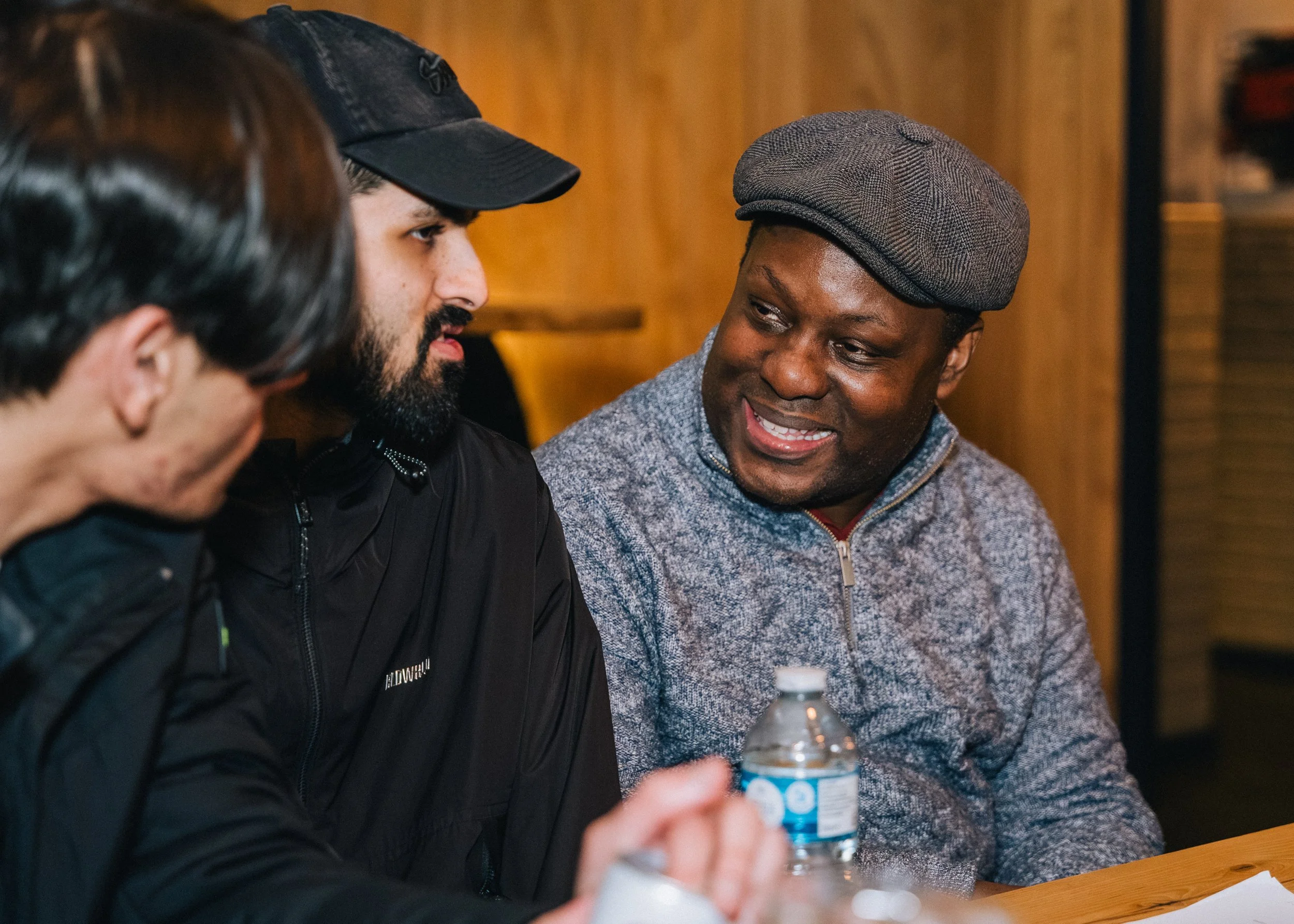 Three men engaged in conversation sitting at a table in a room with wood-paneled walls. One man is wearing a gray cap and sweater, another is wearing a black jacket and cap, and the third has dark hair and glasses. A water bottle is visible on the table.