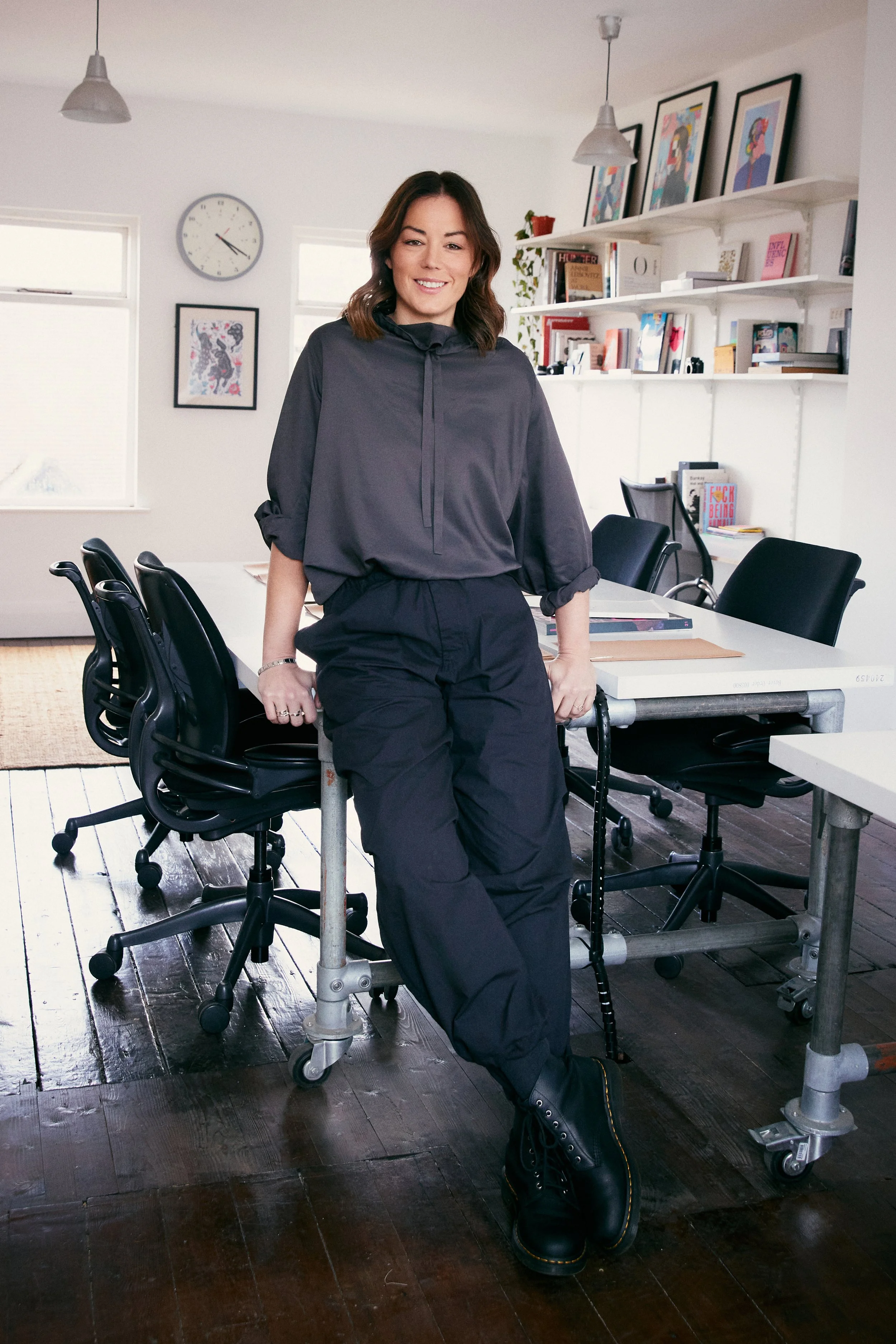 Katy Cowan, a woman with shoulder-length brown hair, smiling, wearing a dark grey jacket and black pants, standing in an office environment with a table and bookshelves in the background.